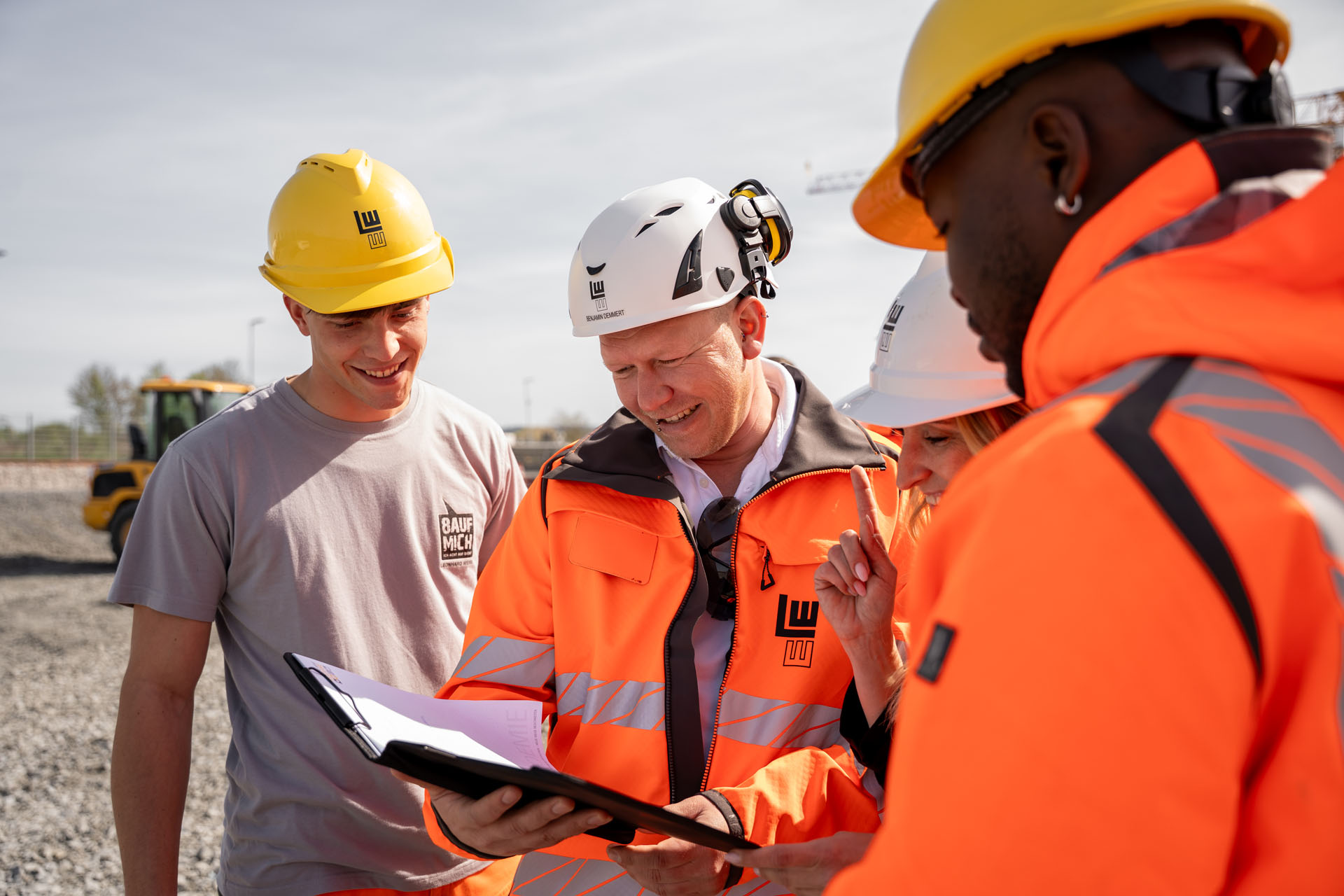 Ein Team von Bauprofis, gekleidet in leuchtend orangefarbene Jacken, analysiert gemeinsam ein Projekt auf einem sonnigen Baustellengelände.