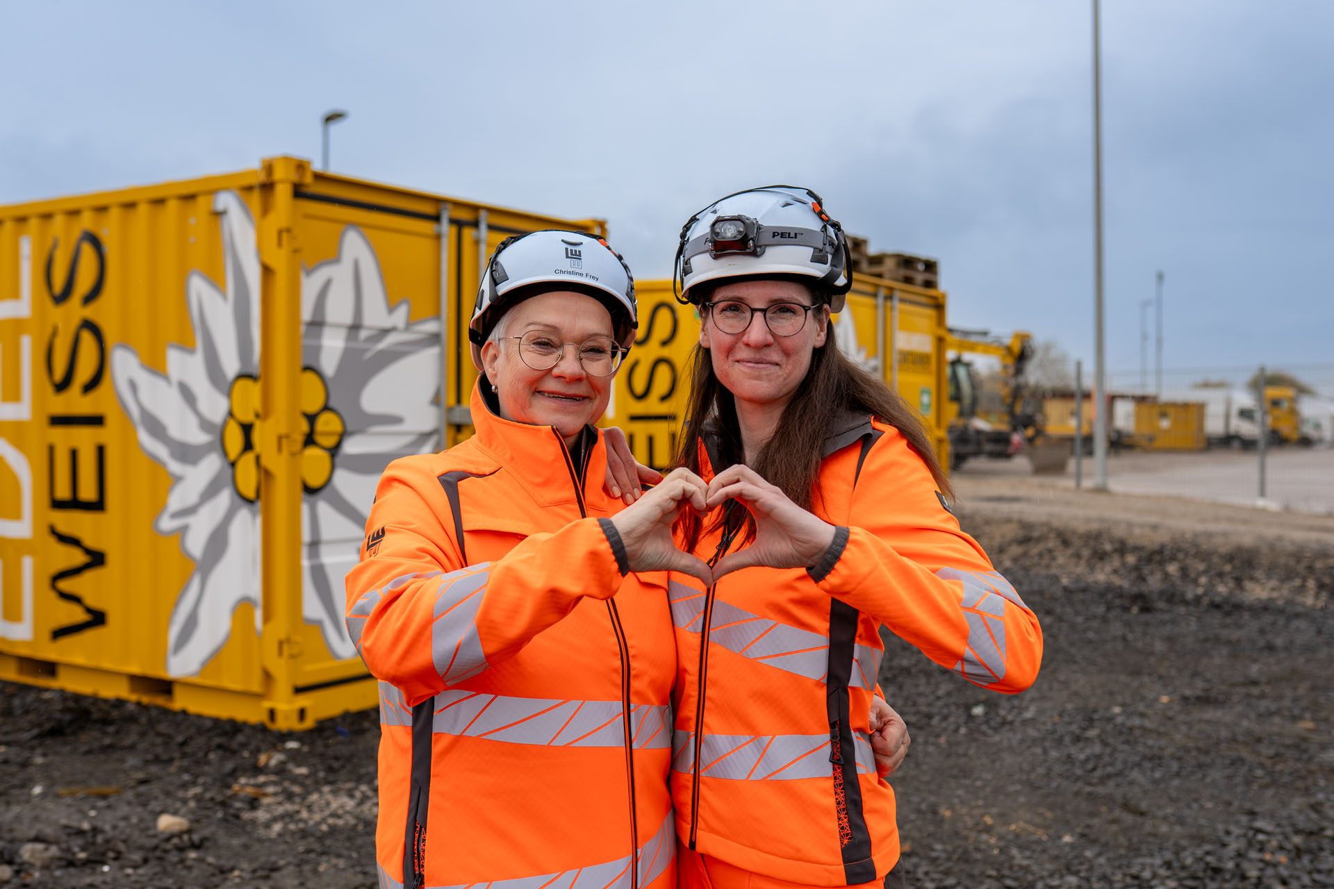Zwei Frauen in leuchtend orangefarbener Arbeitskleidung formen ein Herz mit ihren Händen vor gelben Containern.