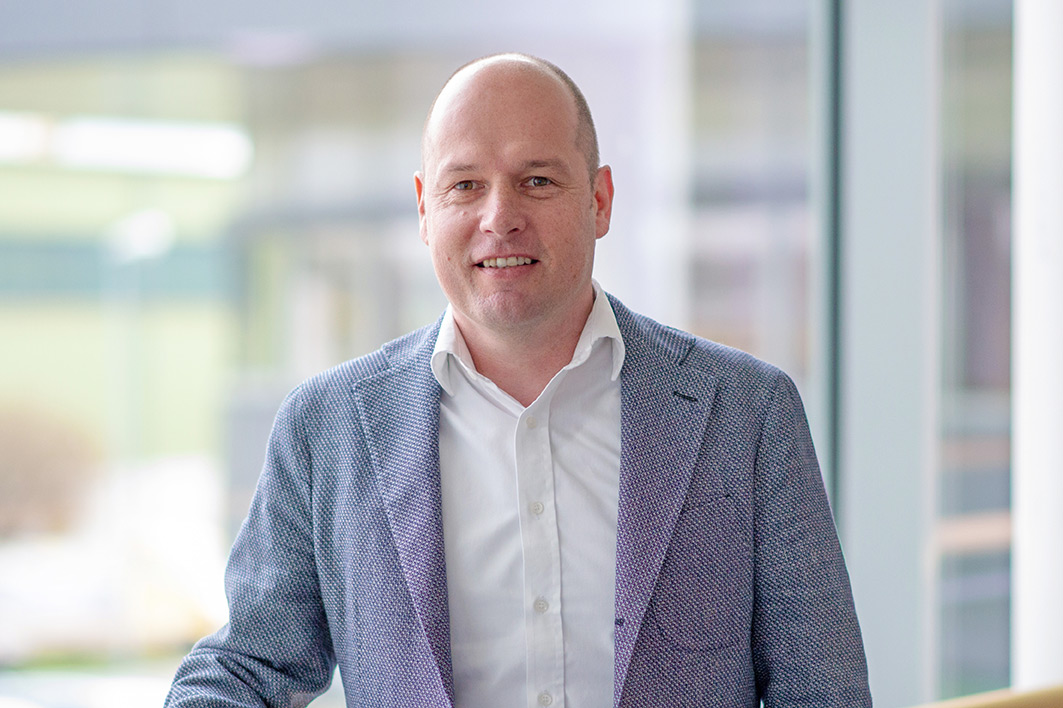 Mit einem warmen Lächeln präsentiert sich ein Mann in einem stilvollen Büro, das von natürlichem Licht durchflutet wird.