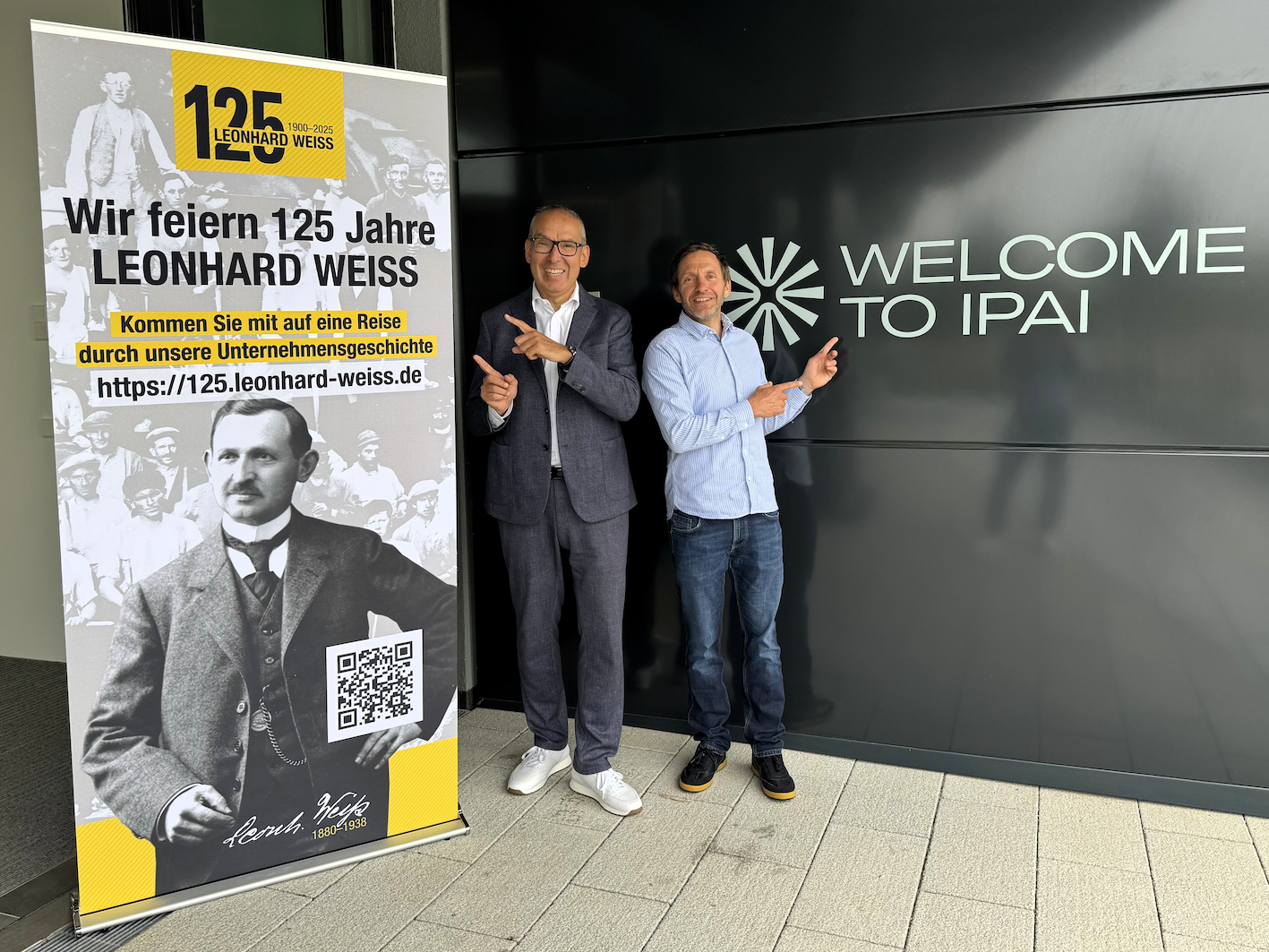 Zwei Männer stehen vor einem Willkommensschild und lächeln in die Kamera.