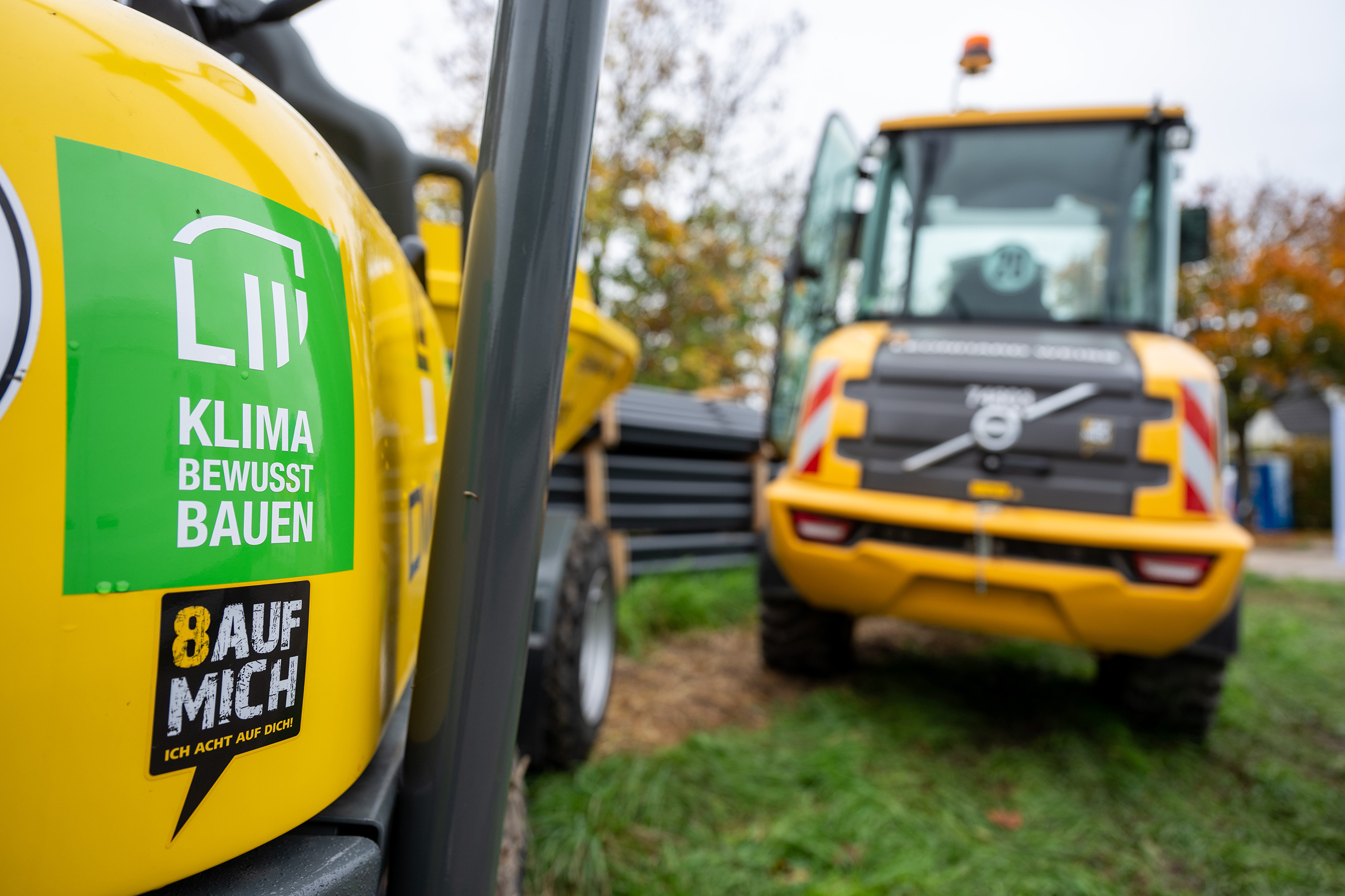 Im Vordergrund ist ein gelbes Baufahrzeug mit dem Focus auf einen Aufkleber, der auf Klimaschutz hinweist, während ein weiteres Fahrzeug im Hintergrund sichtbar ist.