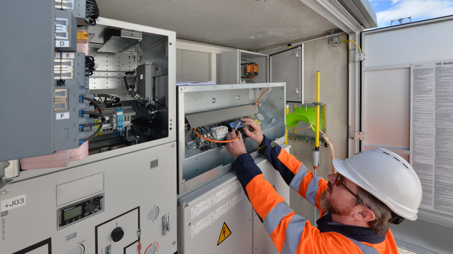 Ein Elektriker mit Schutzhelm und Warnjacke arbeitet an einem offenen Schaltschrank im Freien. Er prüft Kabelverbindungen und Bauteile innerhalb der Anlage. Im Hintergrund sind weitere Schaltschränke, Beschriftungen und ein blauer Himmel sichtbar.