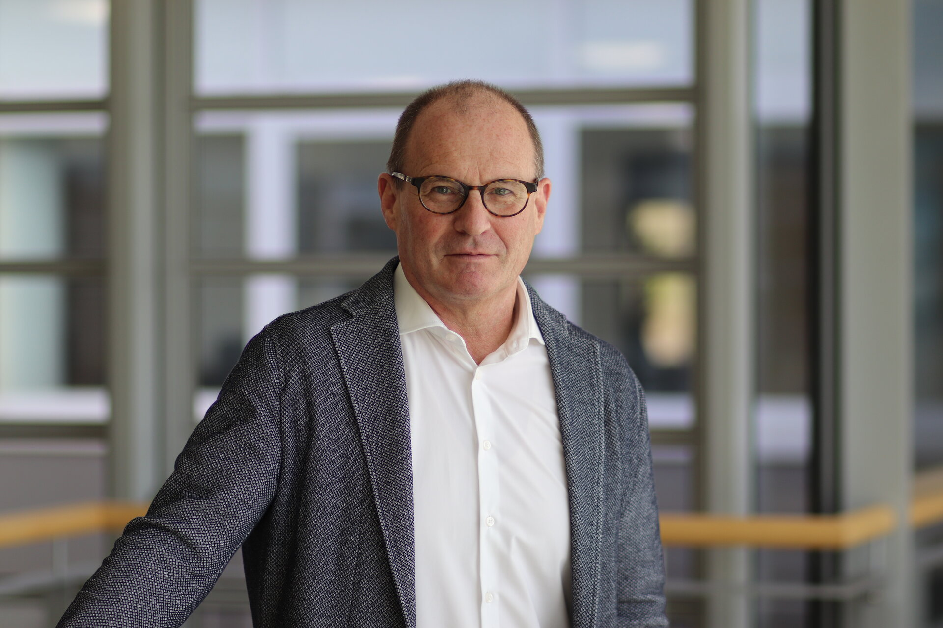 Ein Mann mit Brille steht in einem modernen Raum, lächelt leicht und trägt einen eleganten Anzug mit einem weißen Hemd.