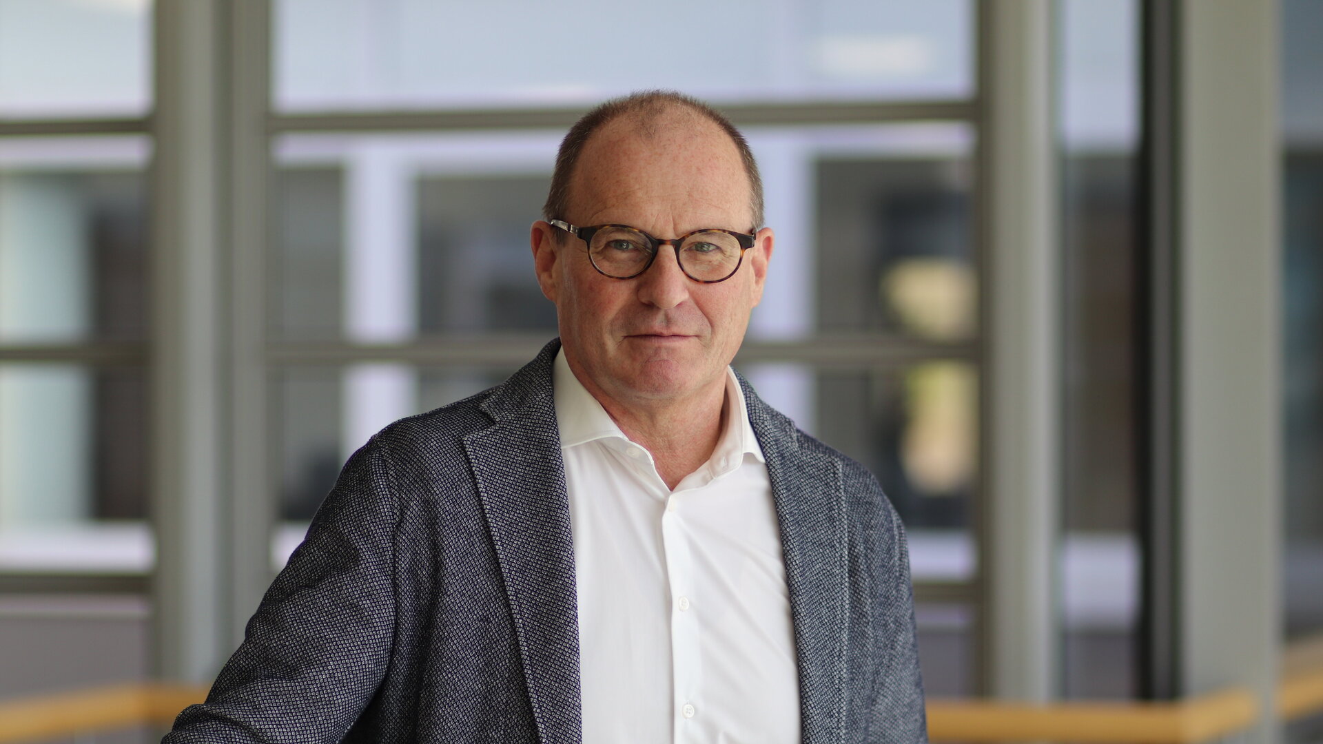 Ein Mann mit Brille steht in einem modernen Raum, lächelt leicht und trägt einen eleganten Anzug mit einem weißen Hemd.