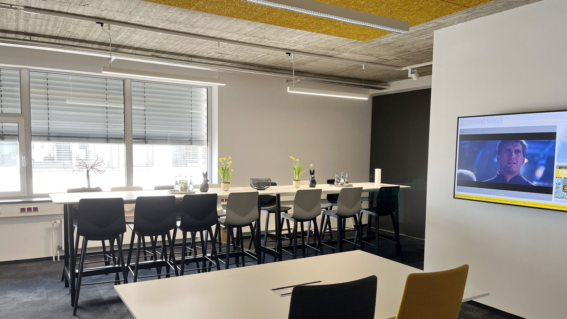 Heller, modern eingerichteter Büro- und Besprechungsraum mit hohen Tischen, Barhockern und einem Bildschirm an der Wand. Auf den Tischen stehen Wasserflaschen und Blumenvasen, die Atmosphäre wirkt offen und einladend.