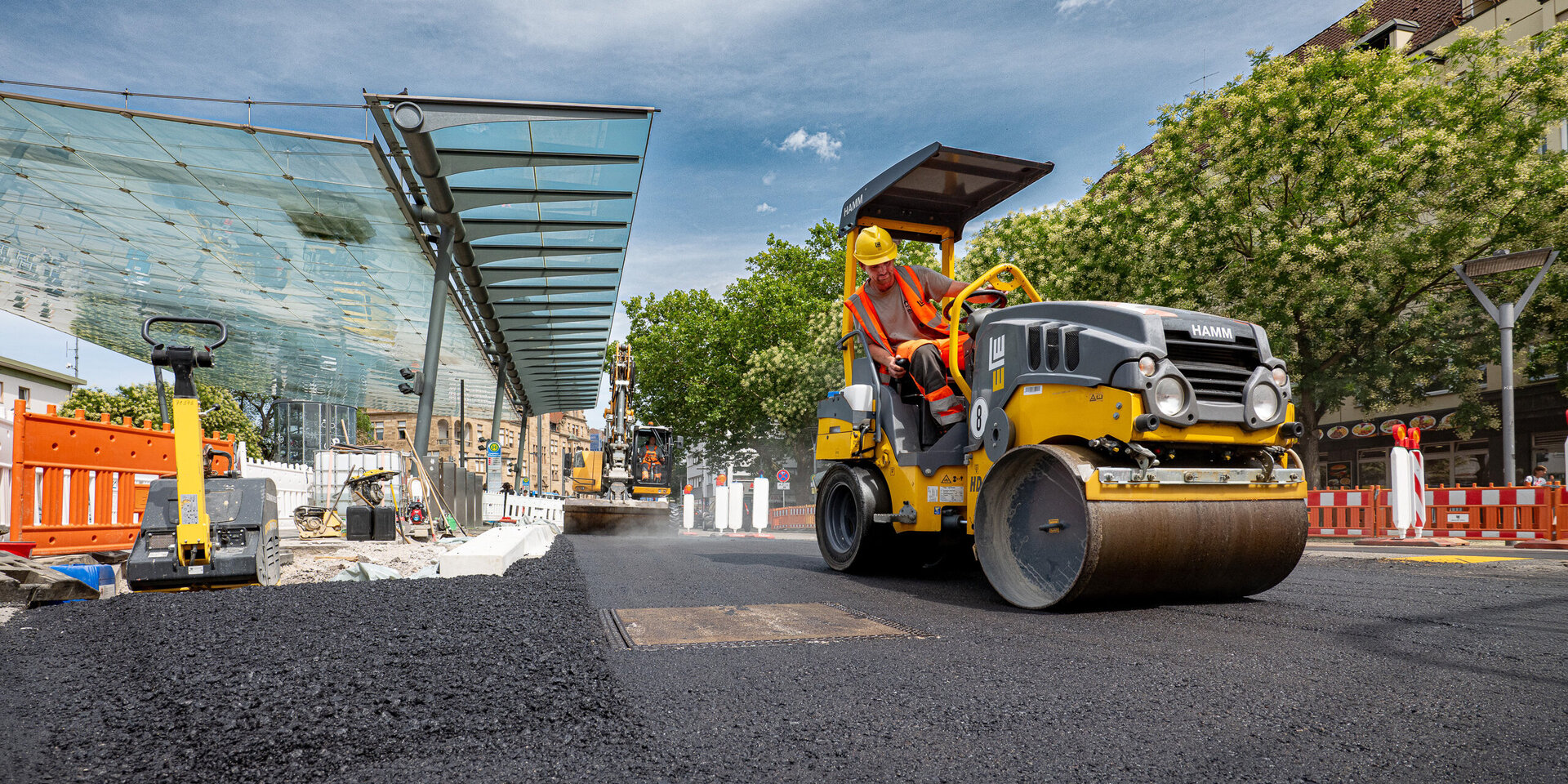 Ein Bauarbeiter in orangefarbener Sicherheitskleidung fährt einen gelben Walzenzug auf einer neu asphaltierten Straße.
