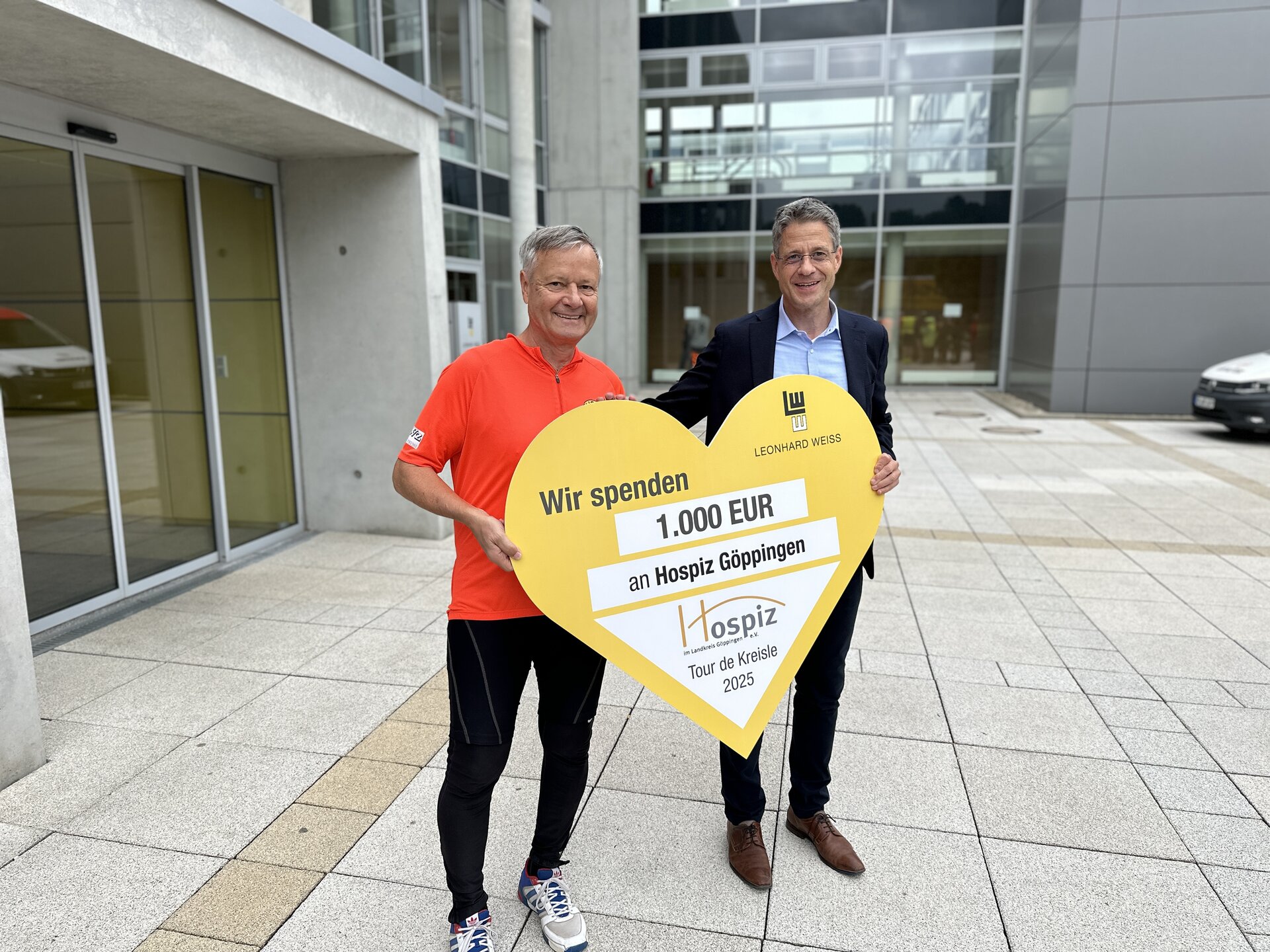 Zwei Männer stehen vor einem modernen Firmengebäude und halten gemeinsam ein großes, gelbes Herz-Schild mit der Aufschrift Wir spenden 1.000 EUR an Hospiz Göppingen – Tour de Kreisel 2025. Der linke Mann trägt Sportkleidung in Rot und Schwarz, der rechte einen Anzug.