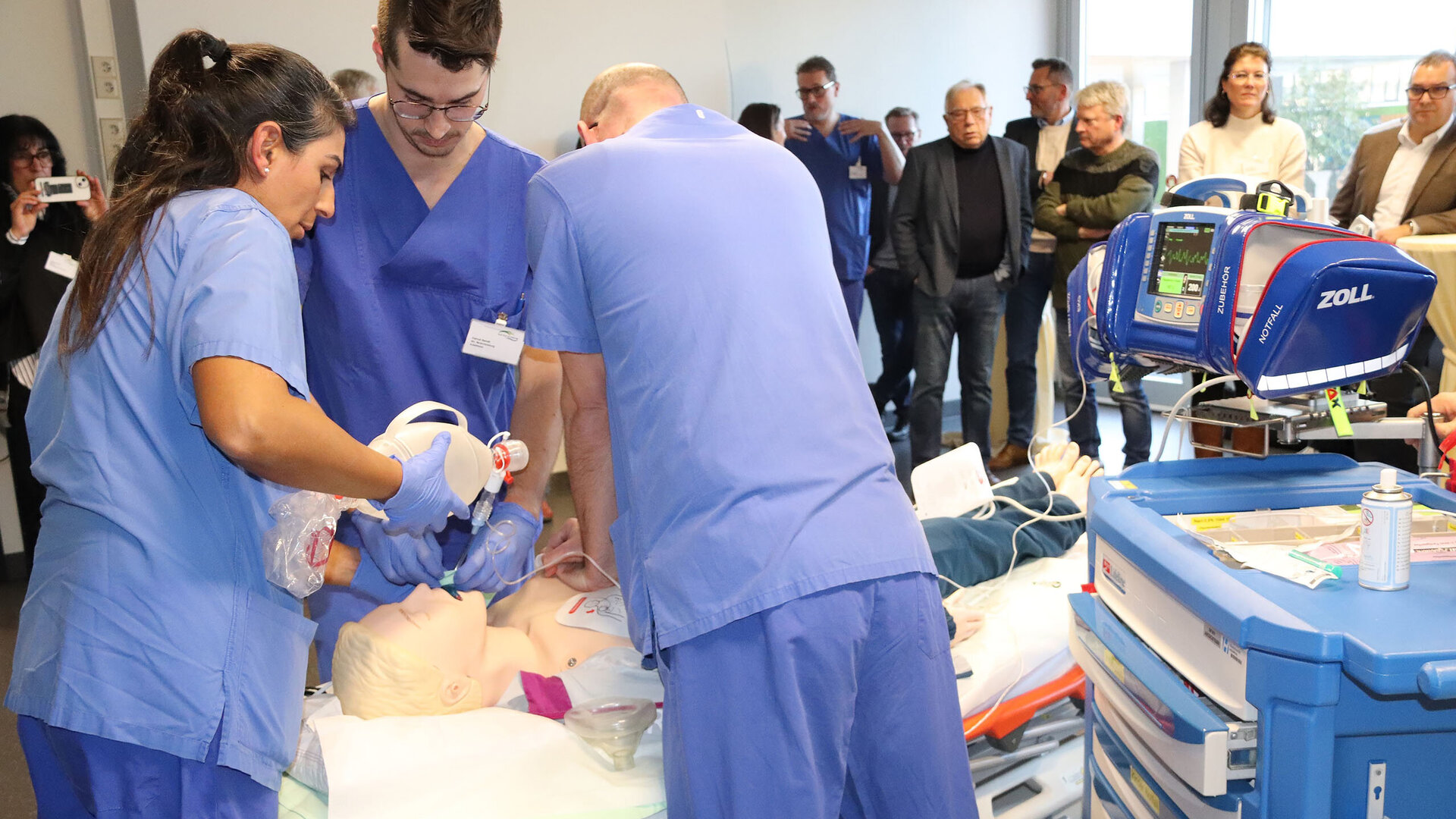 Medizinisches Team in blauen Kitteln führt an einer Reanimationspuppe eine Notfallübung durch, während im Hintergrund eine Gruppe von Zuschauern aufmerksam zusieht.