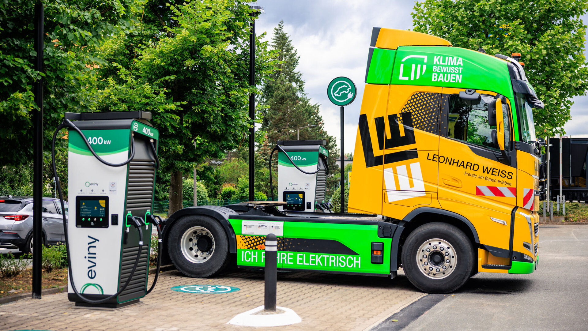 Ein gelber Elektro-LKW von LEONHARD WEISS mit der Aufschrift ‚Ich fahre elektrisch‘ steht an einer 400 kW-Ladesäule. Auf dem Fahrerhaus ist zusätzlich das grüne Logo ‚Klima bewusst bauen‘ zu sehen. Im Hintergrund stehen Bäume, die den Fokus auf Nachhaltigkeit und Umweltfreundlichkeit unterstreichen.