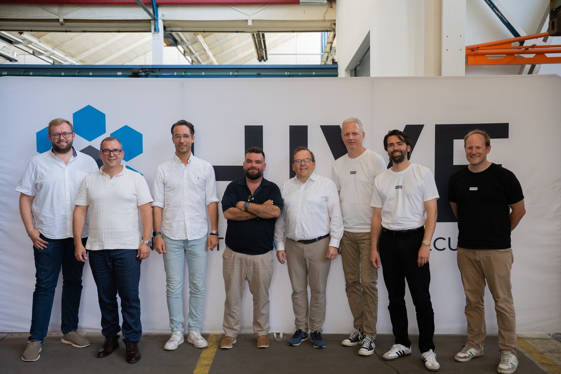 Gruppenfoto beim Richtfest des Projekts HIVE in Göppingen: Acht Männer stehen in einer Reihe vor einer HIVE-Backdrop-Wand in einer Industriehalle. Einige tragen weiße Hemden oder T-Shirts mit HIVE-Logo, andere dunklere Kleidung. Alle blicken lächelnd in die Kamera.