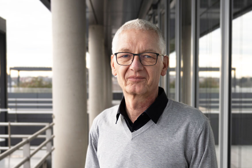 Ein selbstbewusster älterer Herr in einem grauen Pullover steht an einer modernen Gebäudefront mit einem minimalistischen Stil.