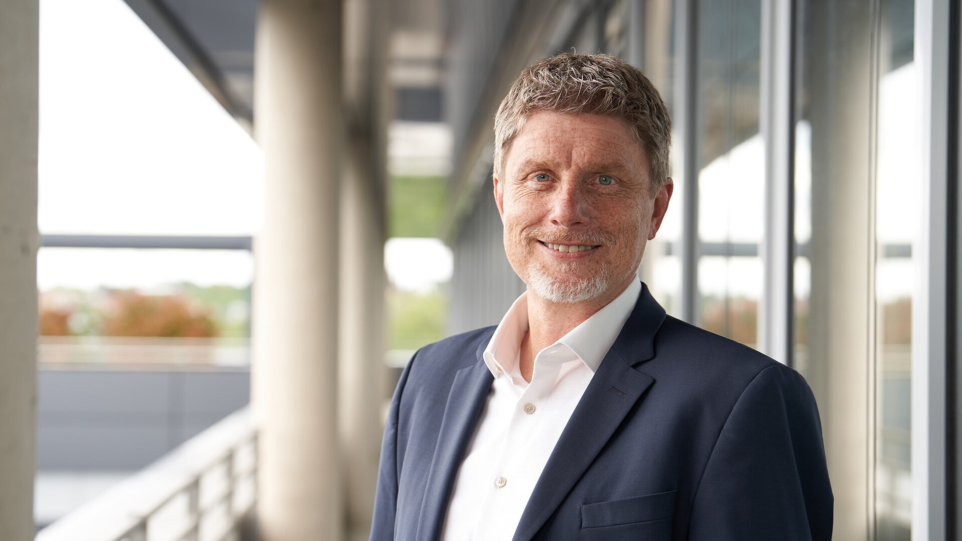 Photo of Robert Kreß, Head of Road and Network Construction, standing in a suit and shirt in front of a modern office building.