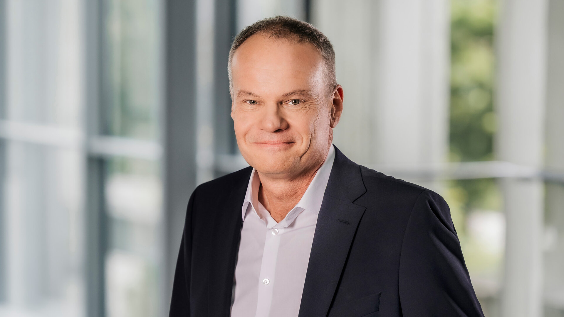 Foto von Christian Ott, Ressortleiter Unternehmenssteuerung und stellvertretender Vorsitzender der Geschäftsführung, stehend in Anzug und Hemd in einem modernen Bürogebäude.