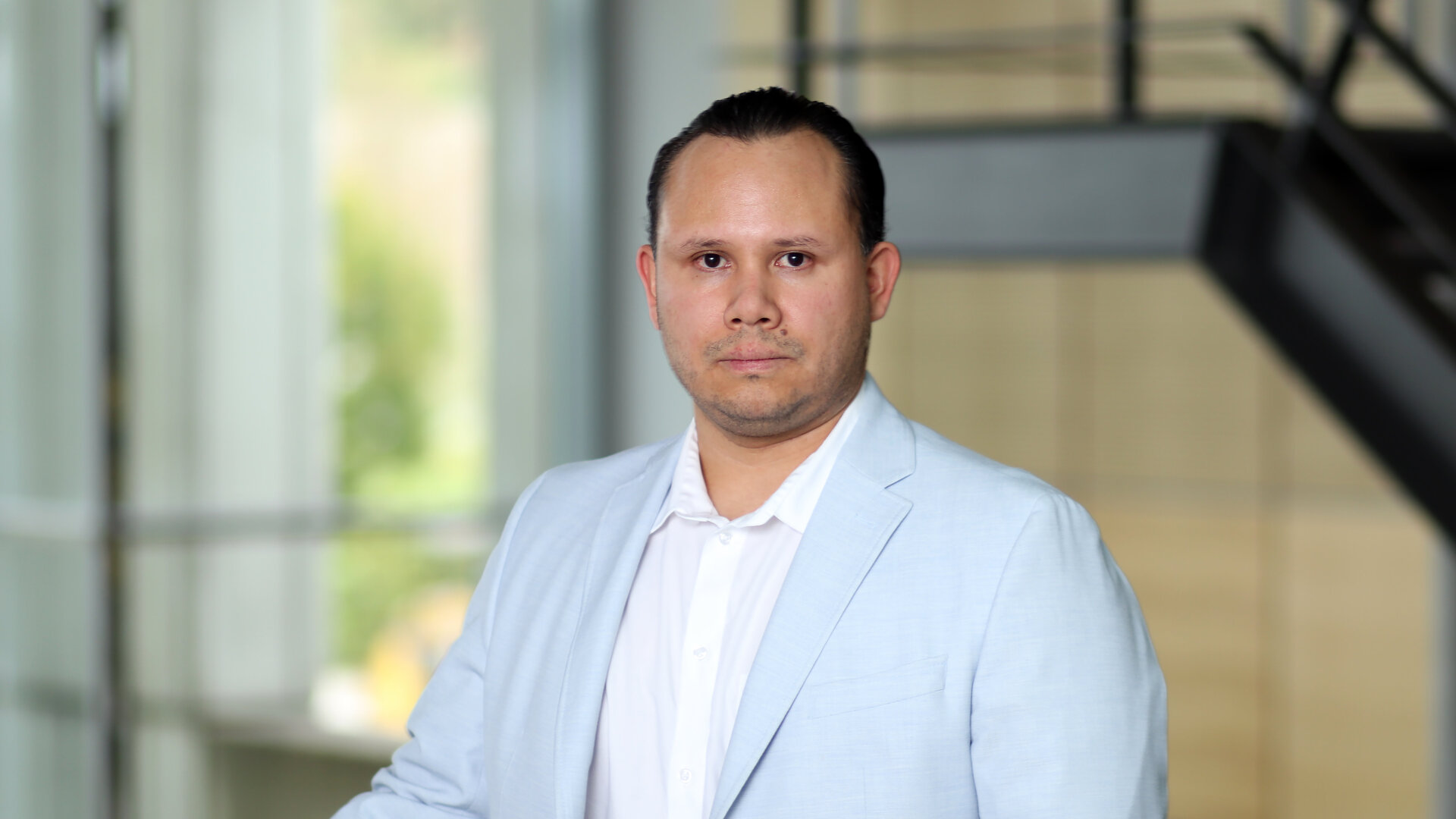 Ein Mann mit einem ernsten Blick trägt ein weißes Hemd unter einem hellblauen Blazer und steht vor einer Glaswand.