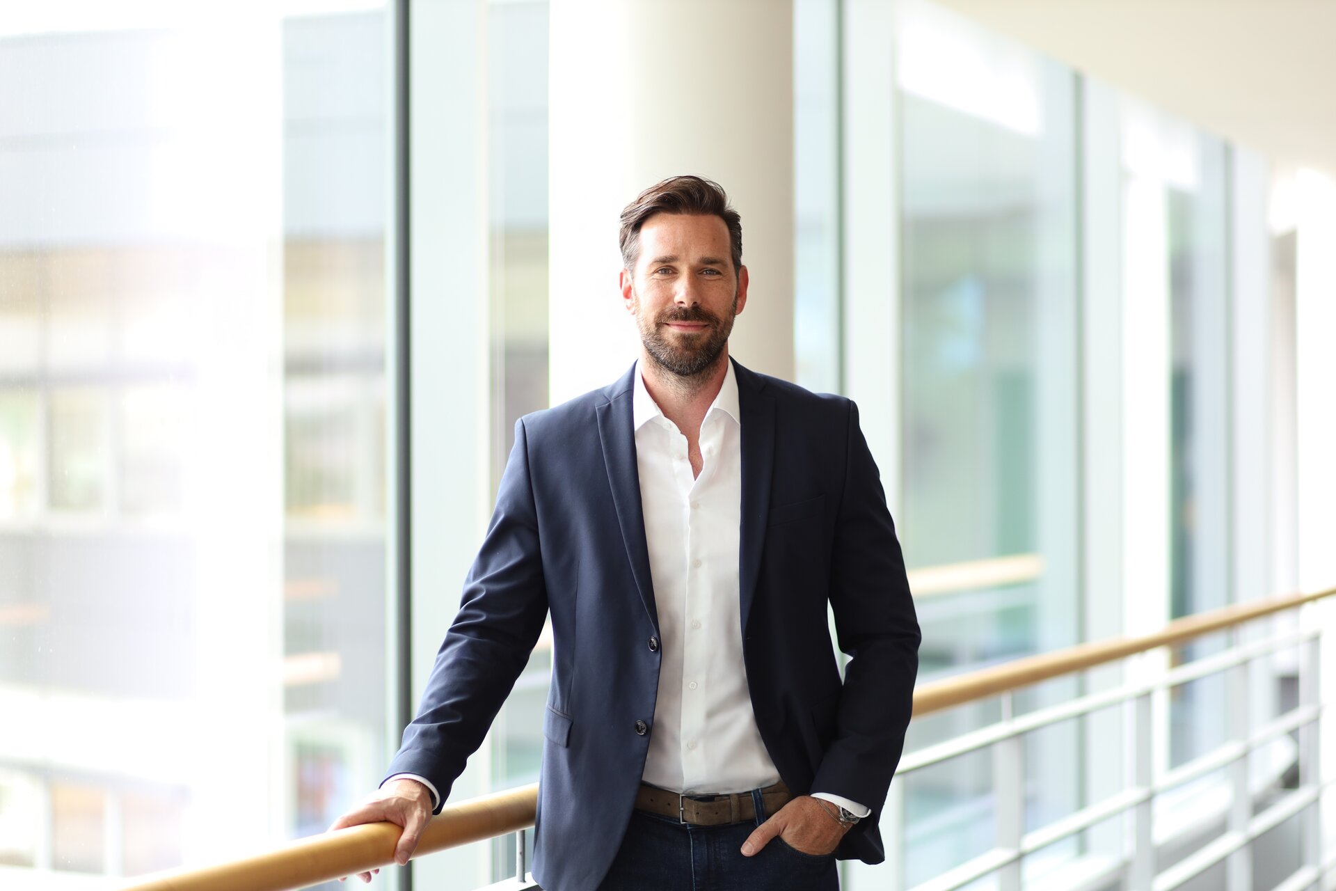 In einem stilvollen Bürogebäude posiert ein Mann in formeller Kleidung vor großen Fenstern mit natürlichem Licht.