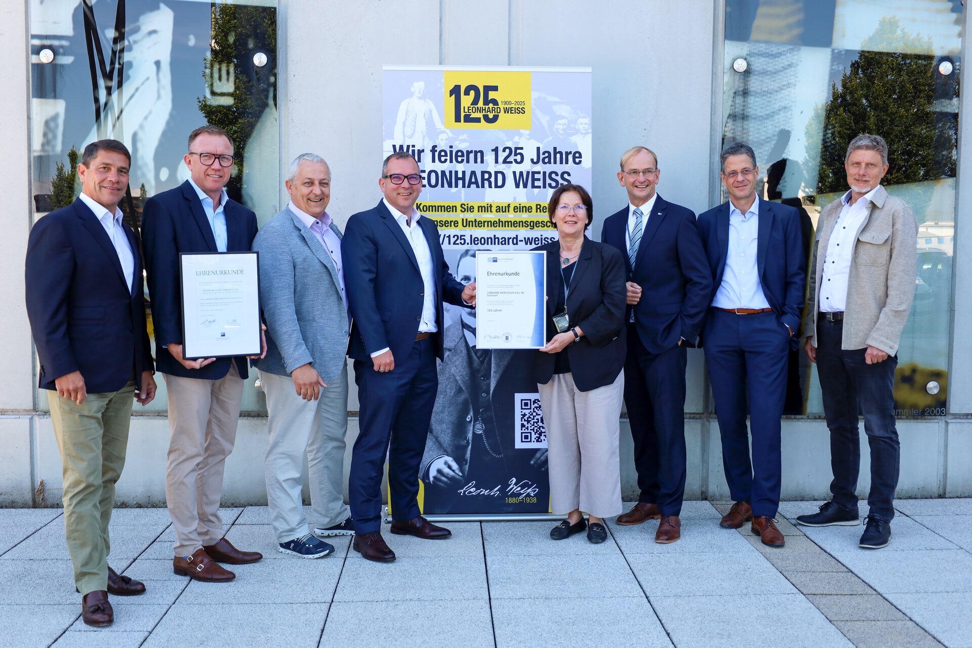 Gruppenfoto vor einem Gebäude mit Banner zum 125-jährigen Jubiläum von Leonhard Weiss. Acht Personen stehen nebeneinander, zwei von ihnen halten gerahmte Ehrenurkunden in den Händen. Im Hintergrund ist ein Jubiläumsplakat mit dem Schriftzug Wir feiern 125 Jahre Leonhard Weiss zu sehen.
