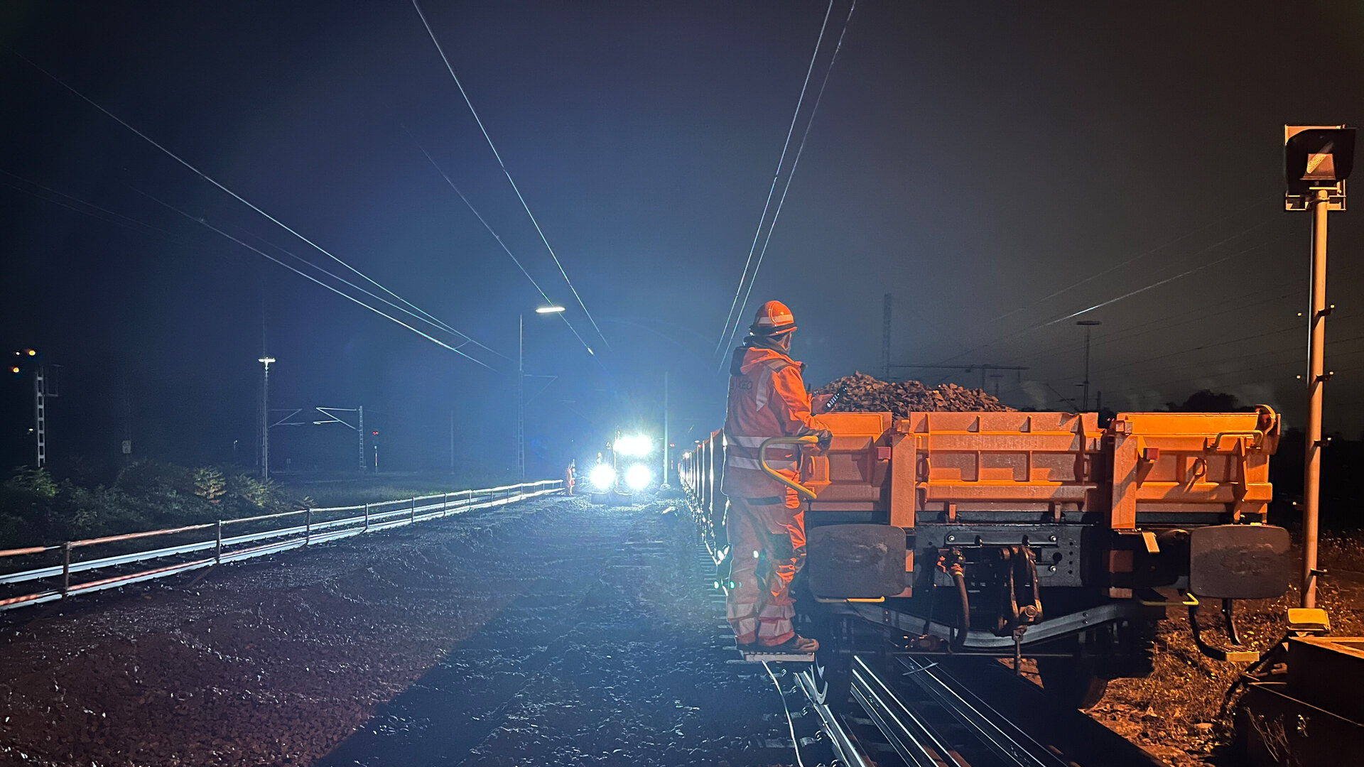 Ein Arbeiter in orangefarbener Sicherheitskleidung steht auf einem Bahnwagen und überwacht das Herstellen von Aushub und das Verladen bei Nacht.