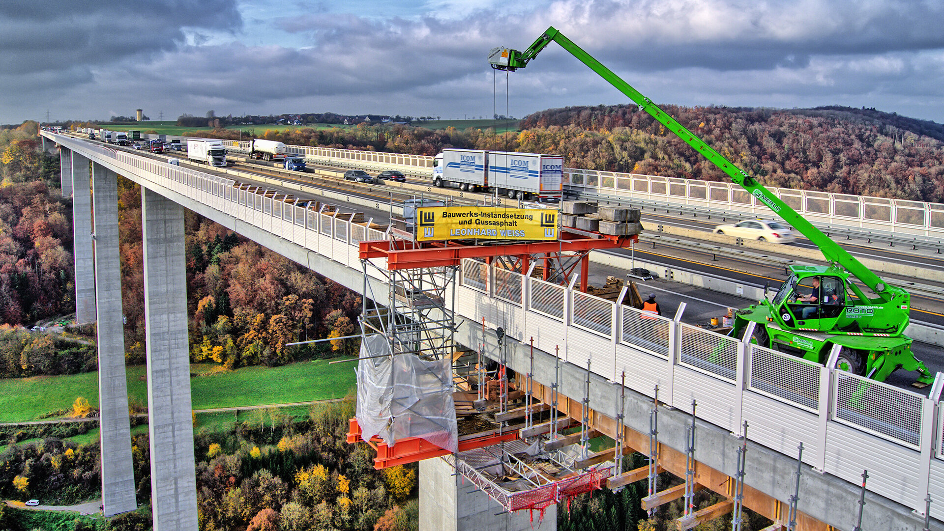 Die Szene zeigt eine Autobahnbrücke mit Bauarbeiten, wo ein grüner Kran Materialien anhebt und die herbstliche Landschaft umrahmt die Szene.