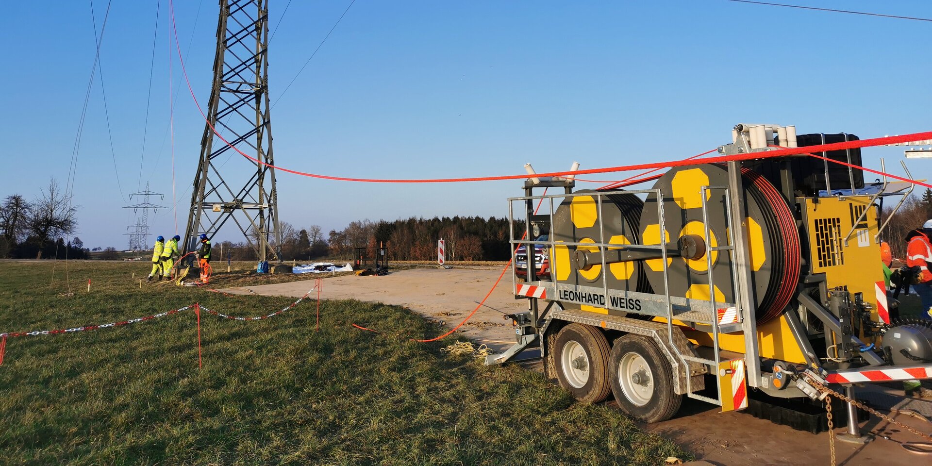 Die Szene zeigt Techniker, die an einem Strommast arbeiten, mit einem gelben Fahrzeug und einem klaren, sonnigen Himmel im Hintergrund.