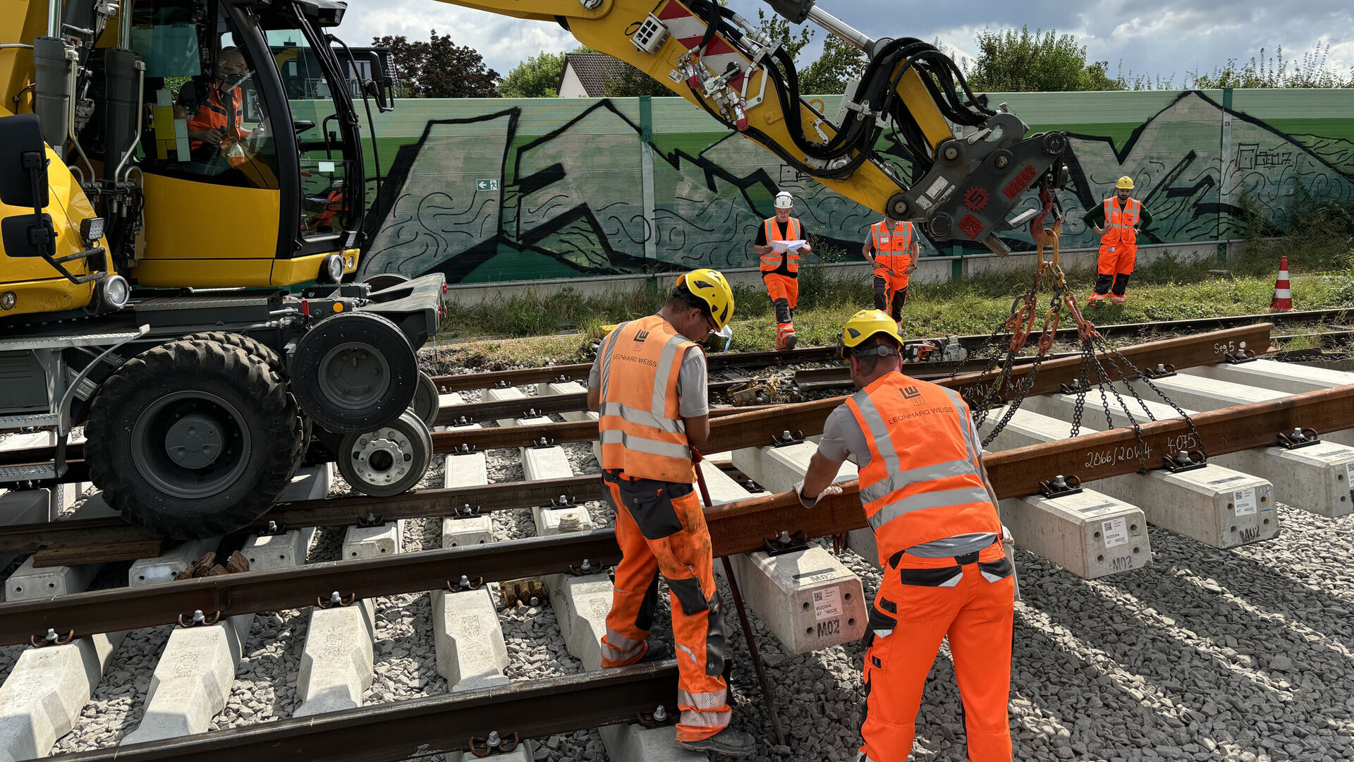 Ein gelber Bagger hebt Schienenstücke an, während Arbeiter in leuchtend orangefarbener Kleidung auf den Gleisen arbeiten.