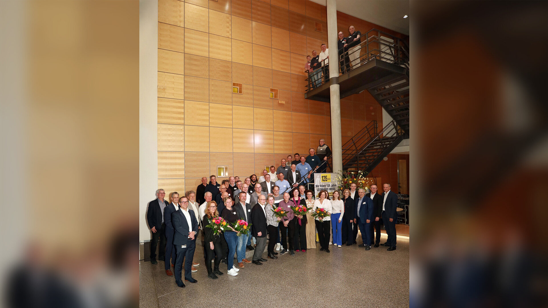 Große Gruppe von Mitarbeitenden und Führungskräften versammelt sich für ein Gruppenfoto in einem modernen Firmengebäude. Einige Personen halten Blumensträuße, im Hintergrund ist ein Banner mit der Aufschrift ‚125 Jahre LEONHARD WEISS‘ zu sehen.