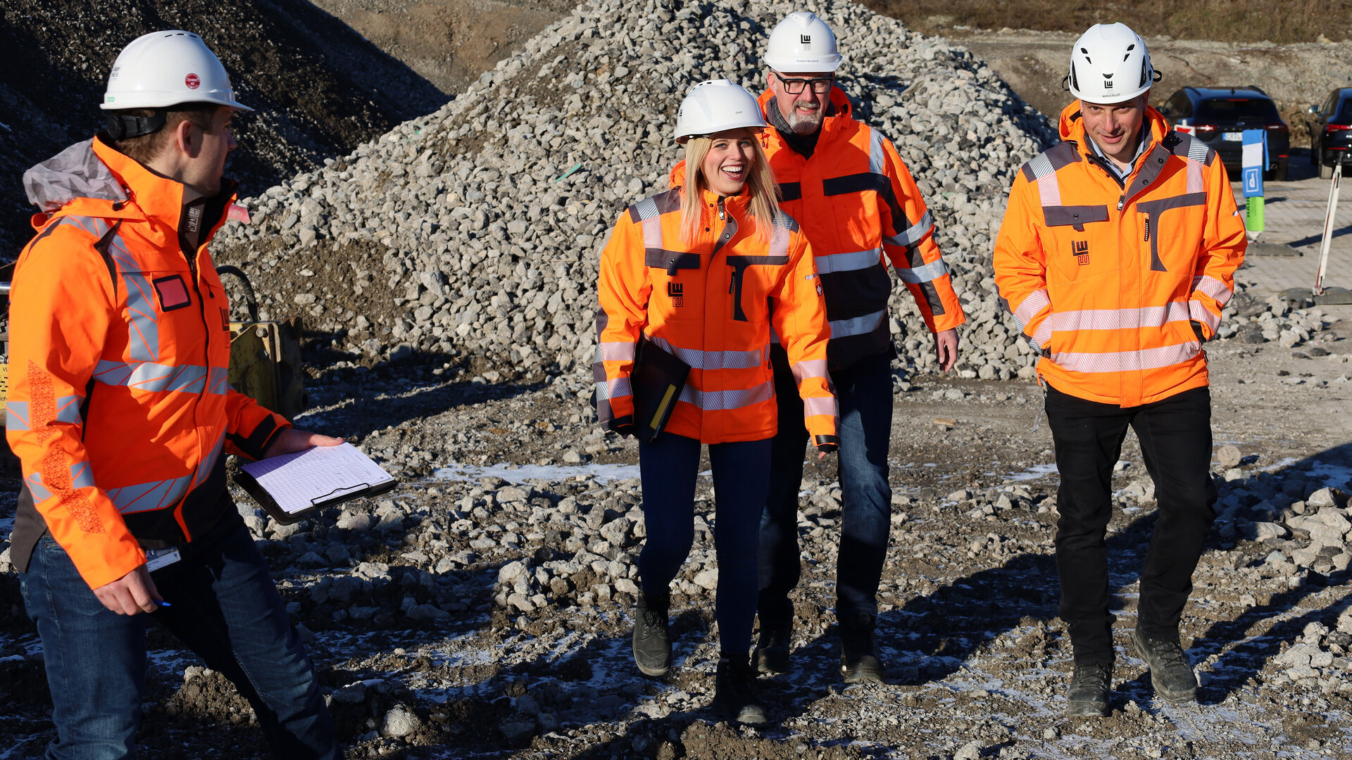 Vier Personen in leuchtend orangefarbener Arbeitskleidung lachen und gehen über eine Baustelle mit Schotterhaufen im Hintergrund.