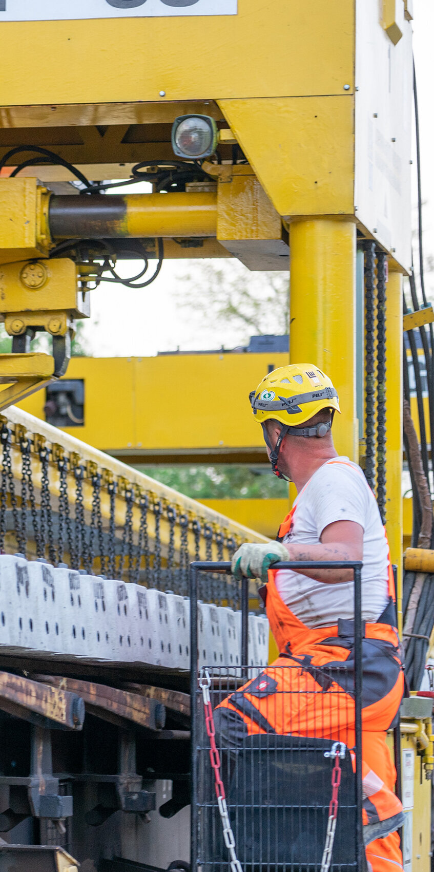 Ein Arbeiter in orangefarbener Arbeitskleidung überwacht den Einsatz eines Krans, der Betonplatten hebt und bewegt.