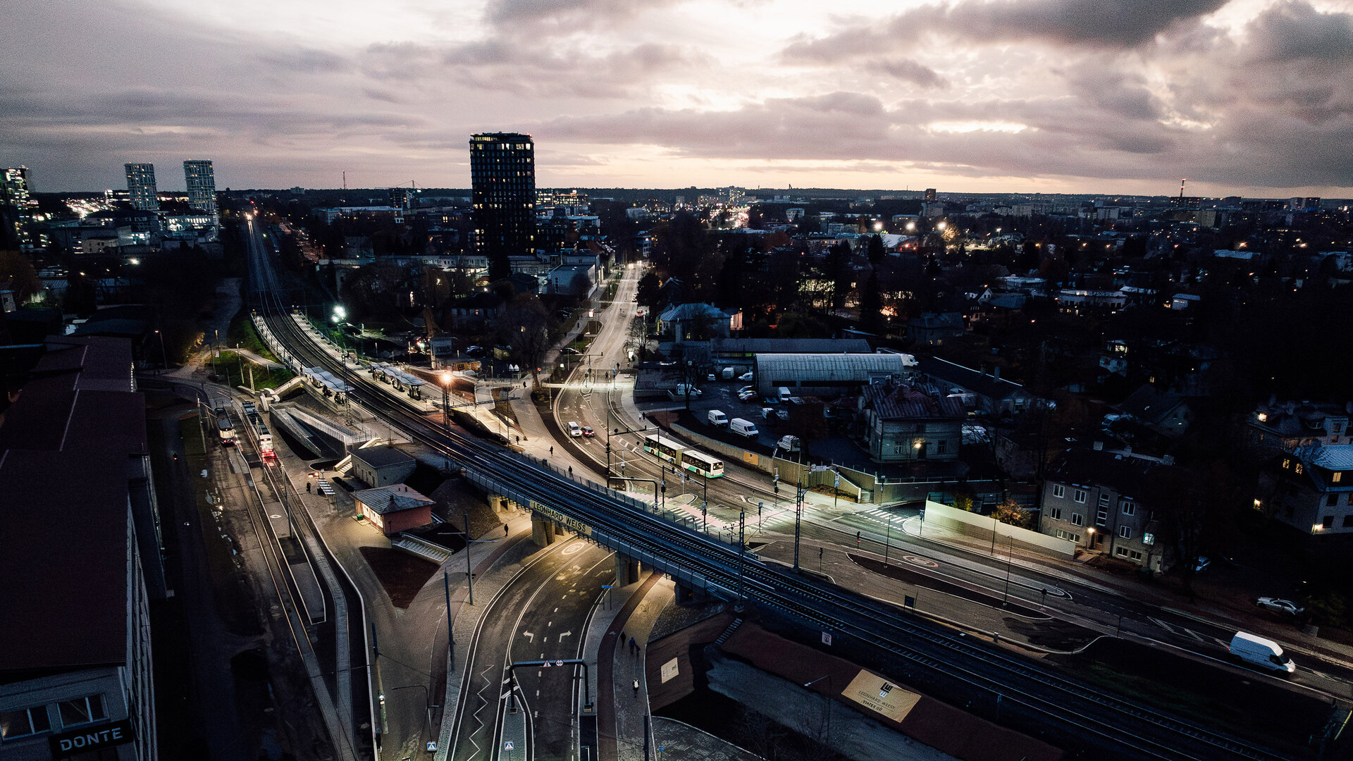Eine weitläufige Stadtansicht mit der Tondi-Kreuzung im Vordergrund bei Dämmerung, mit beleuchteten Straßen und einem komplexen Verkehrsnetz aus Schienen und Straßen.