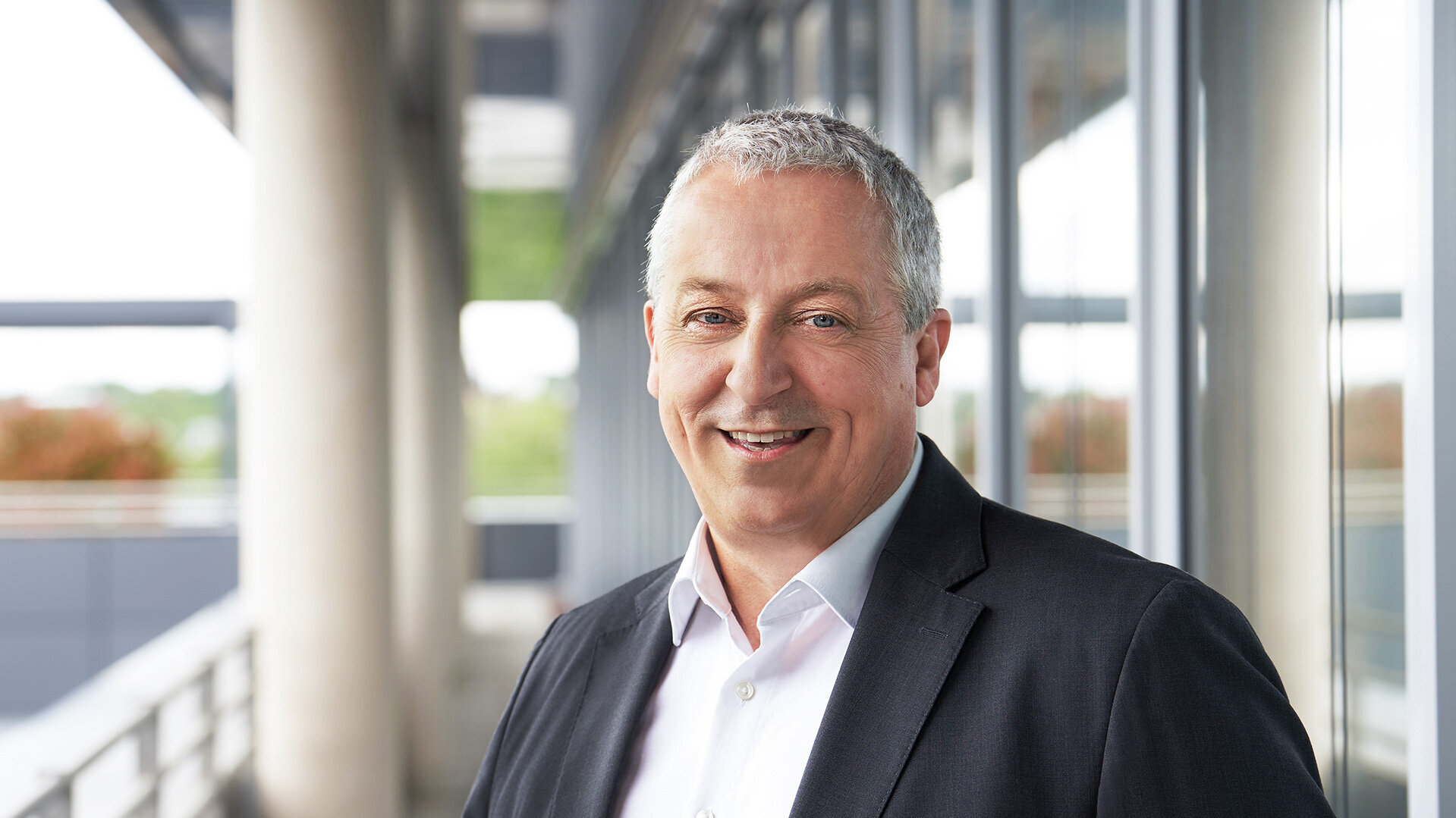 Foto von Ralf Schmidt, Ressortleiter Technik, stehend in Anzug und Hemd vor einem modernen Bürogebäude.