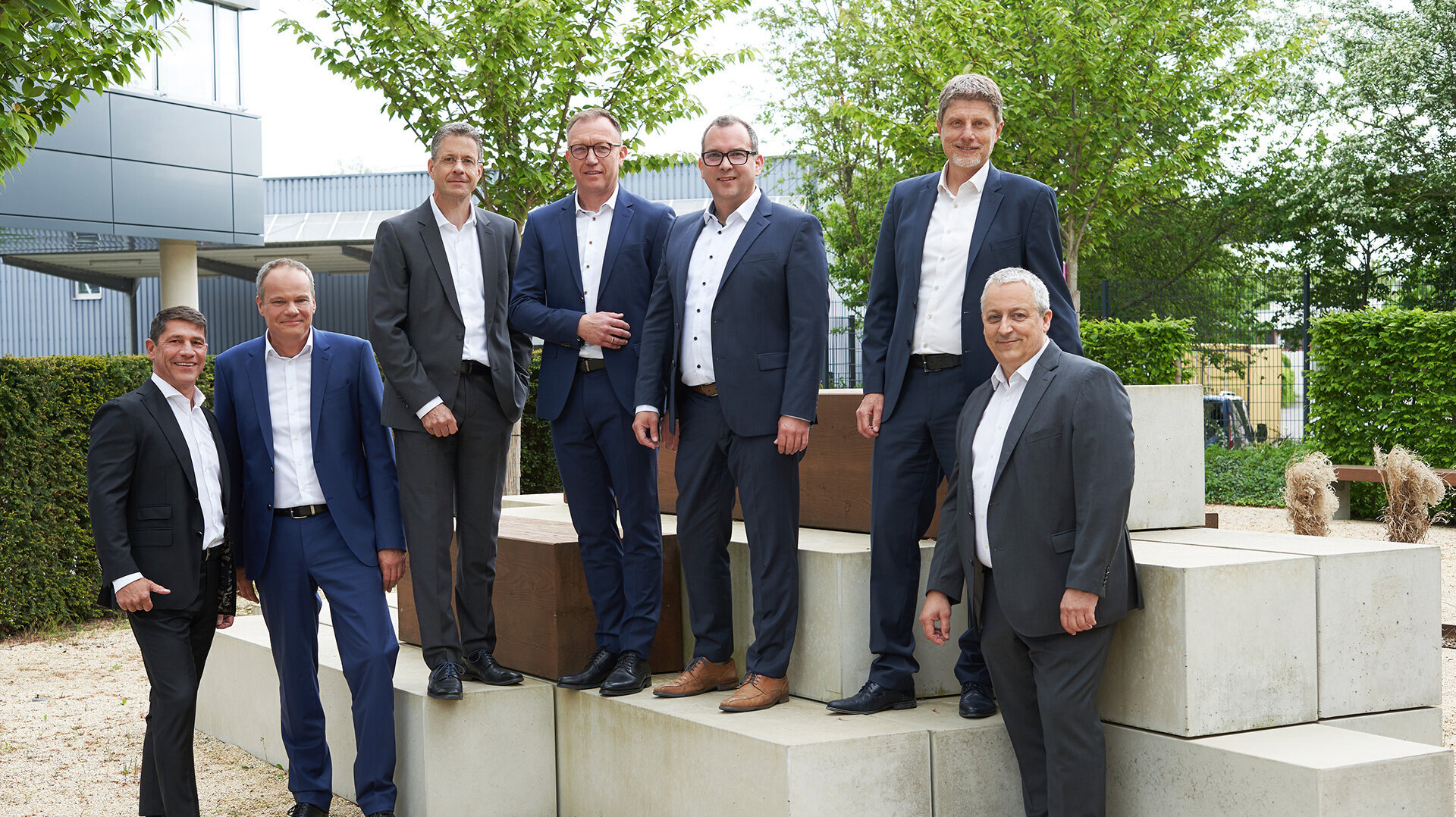 Gruppenbild der Geschäftsführung in Anzügen stehen auf einer Betonstruktur im Freien, umgeben von Bäumen und einem modernen Gebäude.