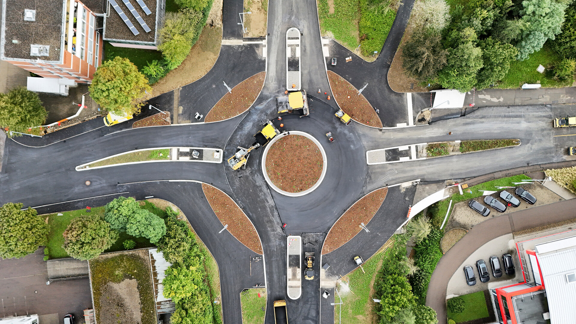 Drohnenaufnahme eines neu gebauten Kreisverkehrs mit frisch asphaltierten Straßen. Mehrere Baustellenfahrzeuge und Walzen sind im Einsatz. Die Mitte des Kreisels ist bepflanzt, umliegende Gebäude, Bäume und ein Parkplatz sind sichtbar.