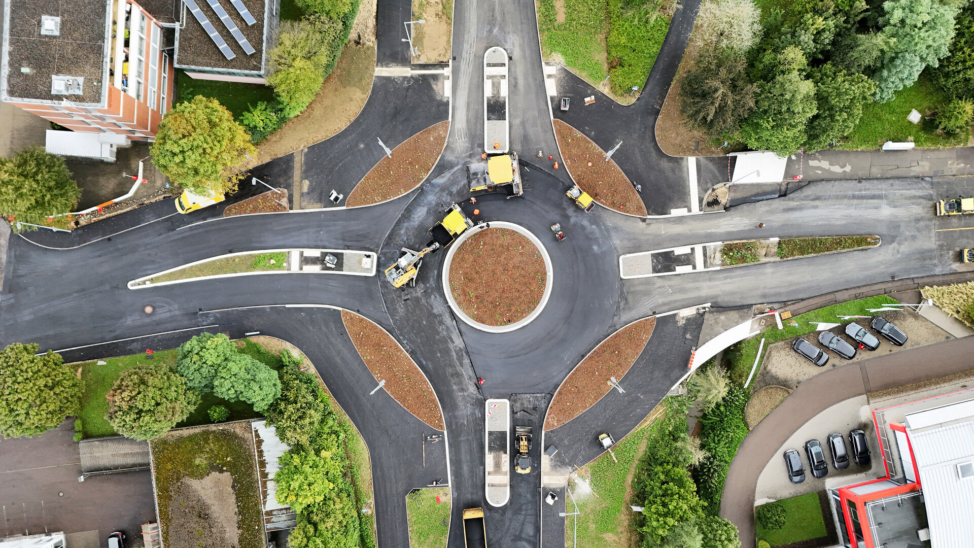 Drohnenaufnahme eines neu gebauten Kreisverkehrs mit frisch asphaltierten Straßen. Mehrere Baustellenfahrzeuge und Walzen sind im Einsatz. Die Mitte des Kreisels ist bepflanzt, umliegende Gebäude, Bäume und ein Parkplatz sind sichtbar.