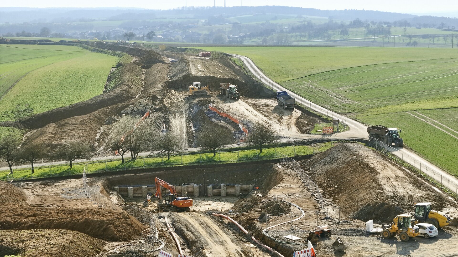 Eine Baustelle mit schweren Maschinen, die Erde bewegen, umgeben von grünen Feldern und einem klaren Himmel im Hintergrund.