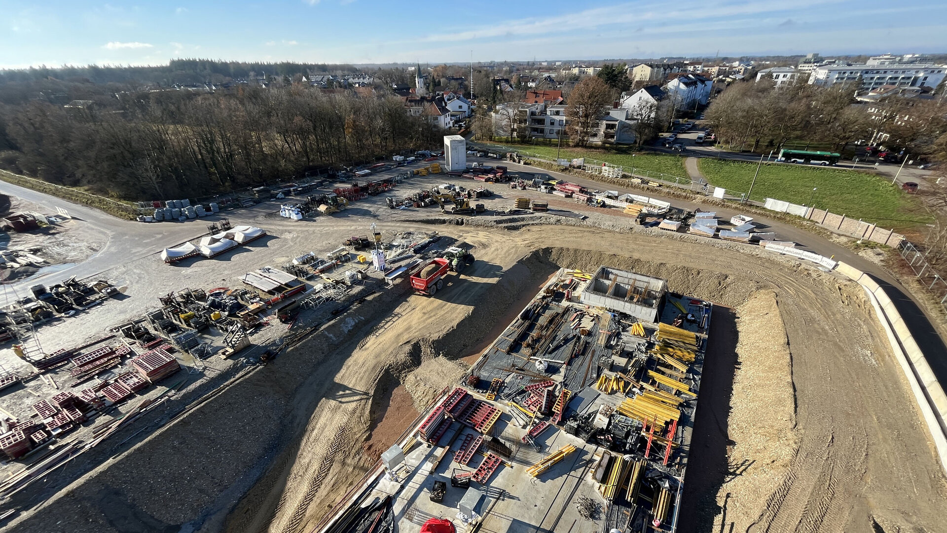 Ein Baustellenbereich mit zahlreichen Baustellenfahrzeugen und -materialien, umgeben von einer grünen Landschaft und Wohnhäusern.