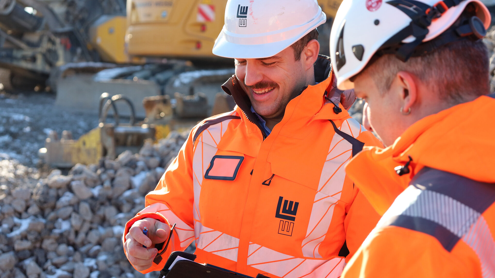 Zwei Bauarbeiter in orangefarbener Schutzkleidung und weißen Helmen im Gespräch auf einer Baustelle. Einer von ihnen hält ein Klemmbrett und lächelt, im Hintergrund sind Baumaschinen und Schotter zu sehen.