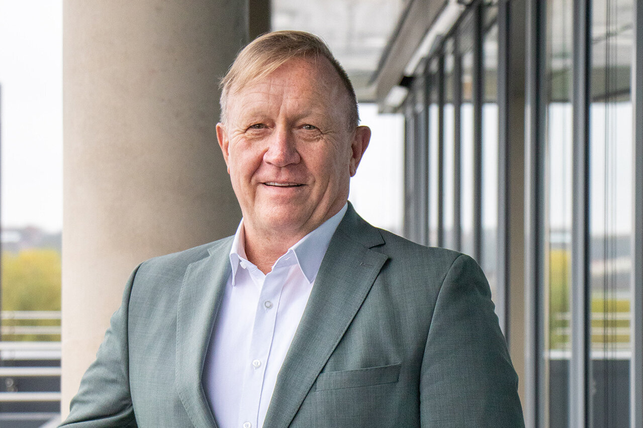 Vor einer modernen Glasfassade steht ein Mann, der in einem grauen Blazer und einem weißen Hemd gekleidet ist.