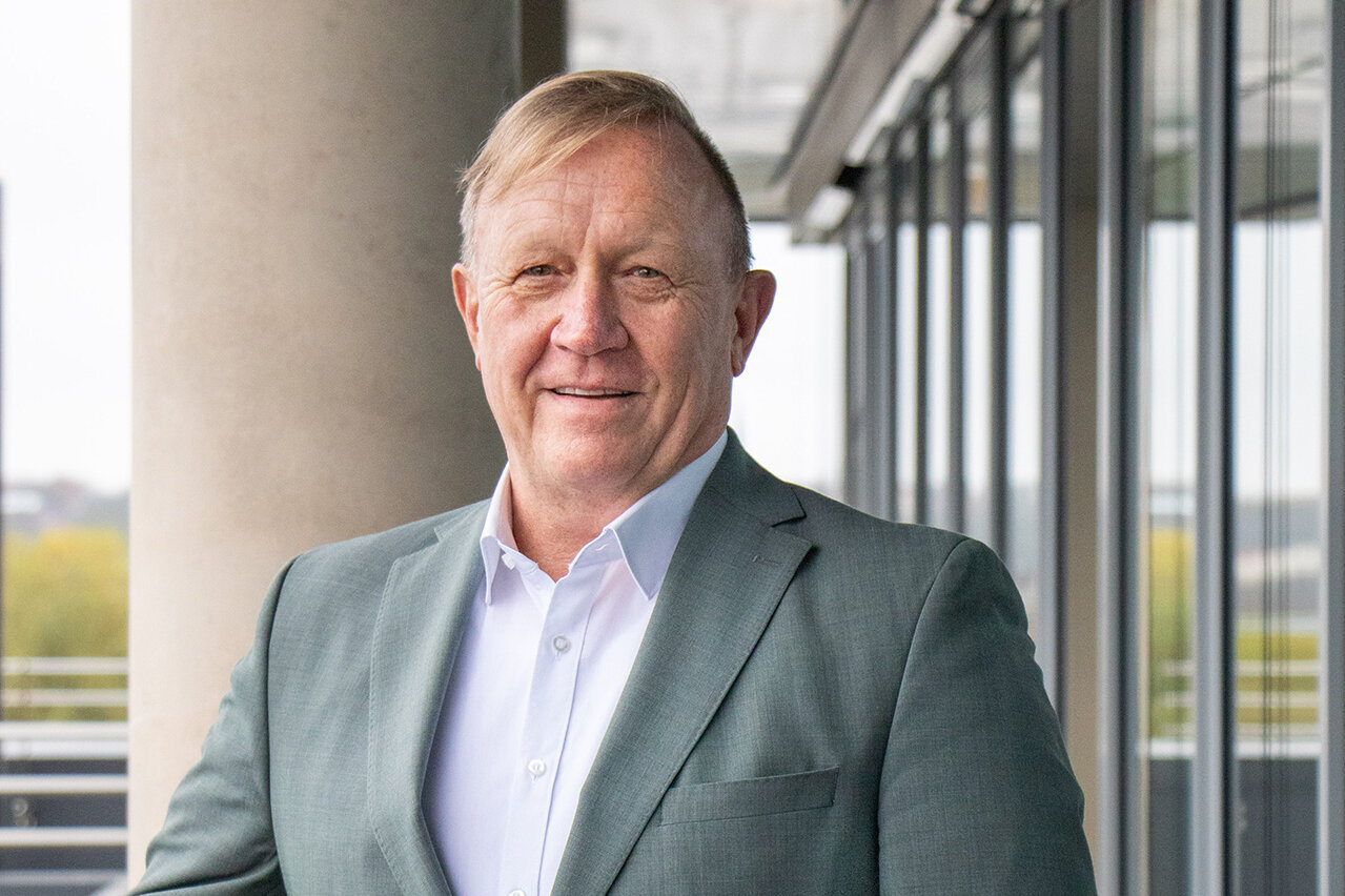 Vor einer modernen Glasfassade steht ein Mann, der in einem grauen Blazer und einem weißen Hemd gekleidet ist.