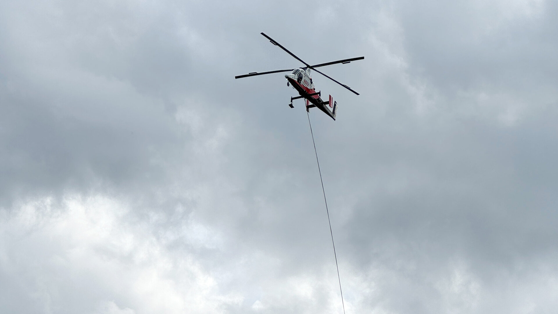 Ein Hubschrauber schwebt über einem Wald und transportiert einen Stahlmasten für die Errichtung einer Lärmschutzwand.