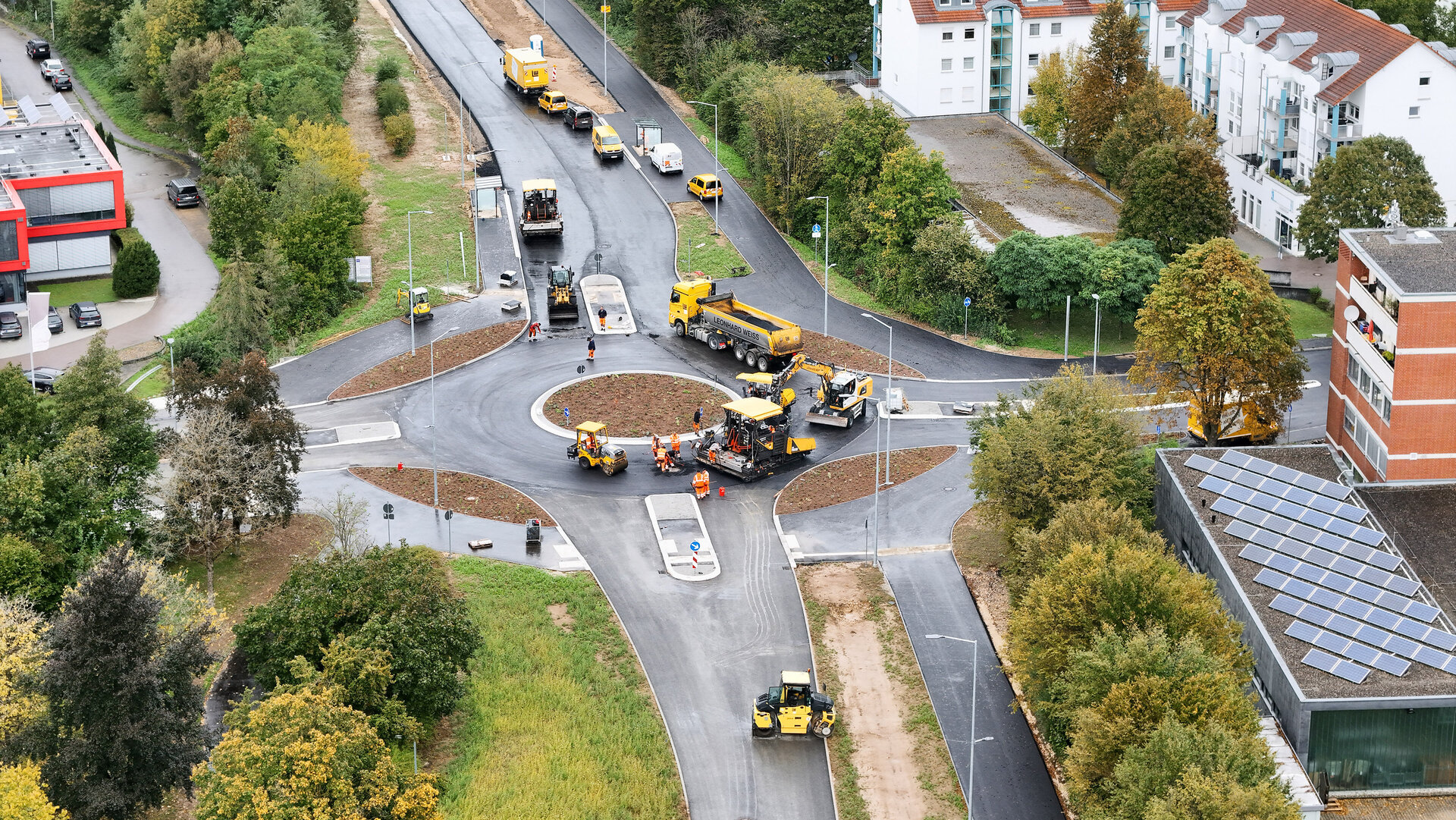 Eine Baustelle mit mehreren gelben Baumaschinen, die an einem neuen Kreisverkehr arbeiten, umgeben von Bäumen und Wohngebäuden.