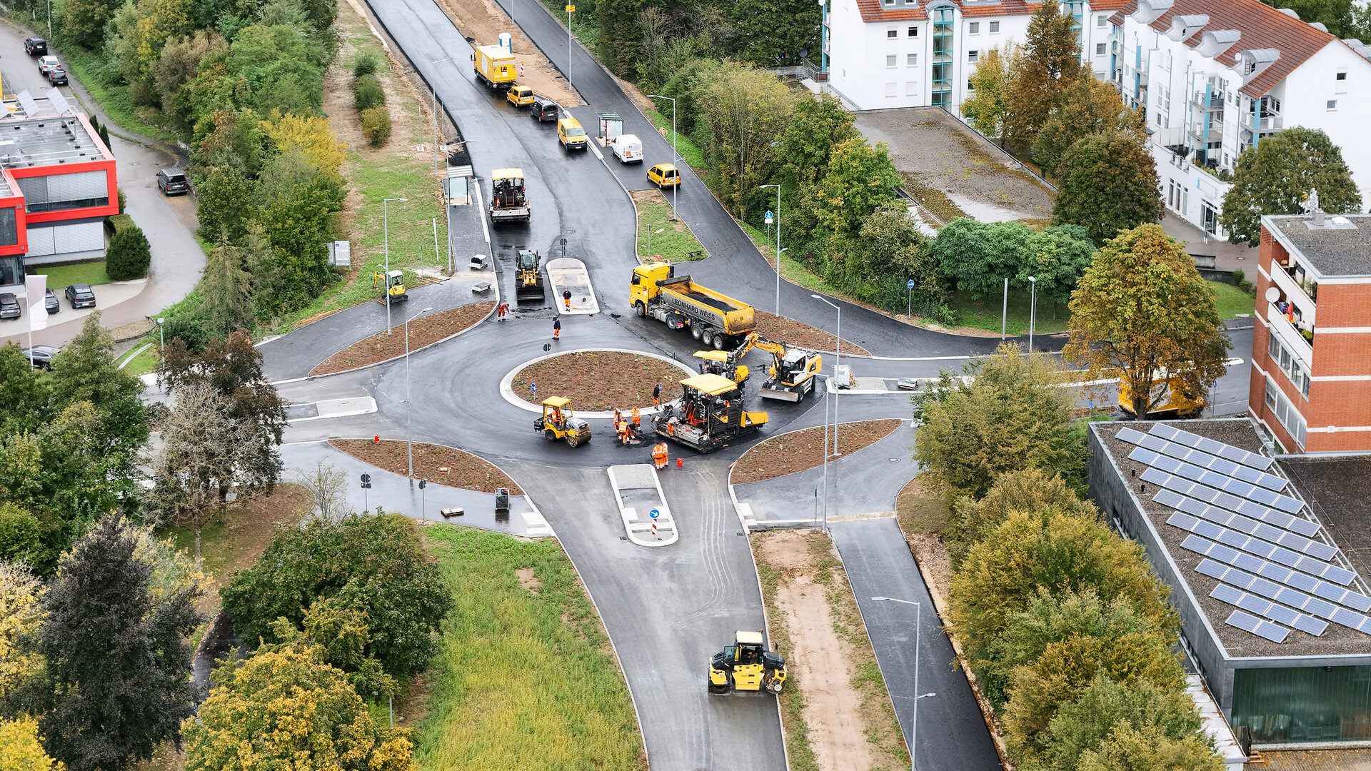 Eine Baustelle mit mehreren gelben Baumaschinen, die an einem neuen Kreisverkehr arbeiten, umgeben von Bäumen und Wohngebäuden.