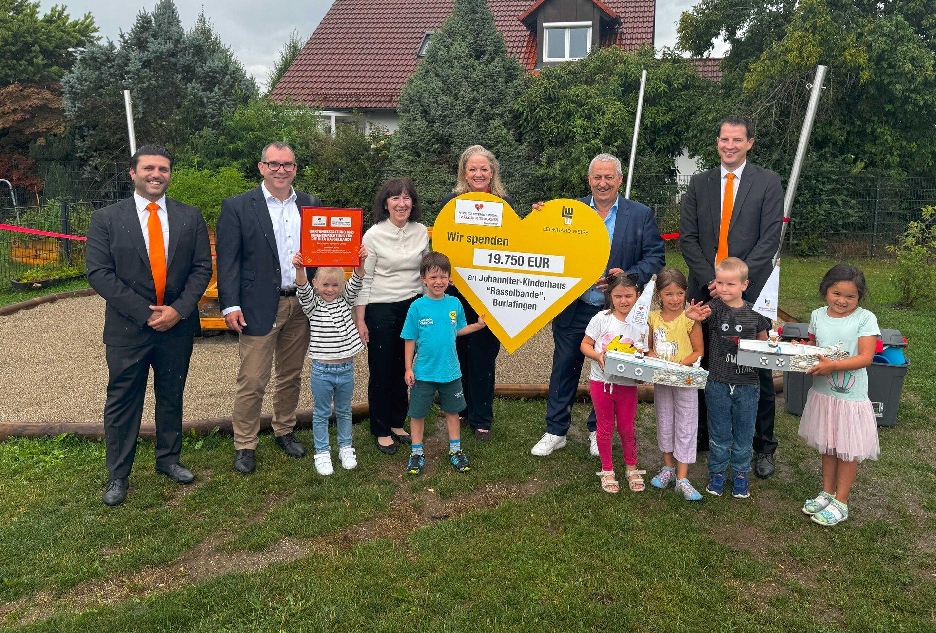 Spendenübergabe an das Johanniter-Kinderhaus ‚Rasselbande‘ in Burlafingen: Vertreter von Leonhard Weiss übergeben gemeinsam mit Kindern und Betreuern ein großes gelbes Herz mit der Spendensumme von 19.750 Euro. Die Kinder halten stolz Schilder und gebastelte Schiffe in den Händen.