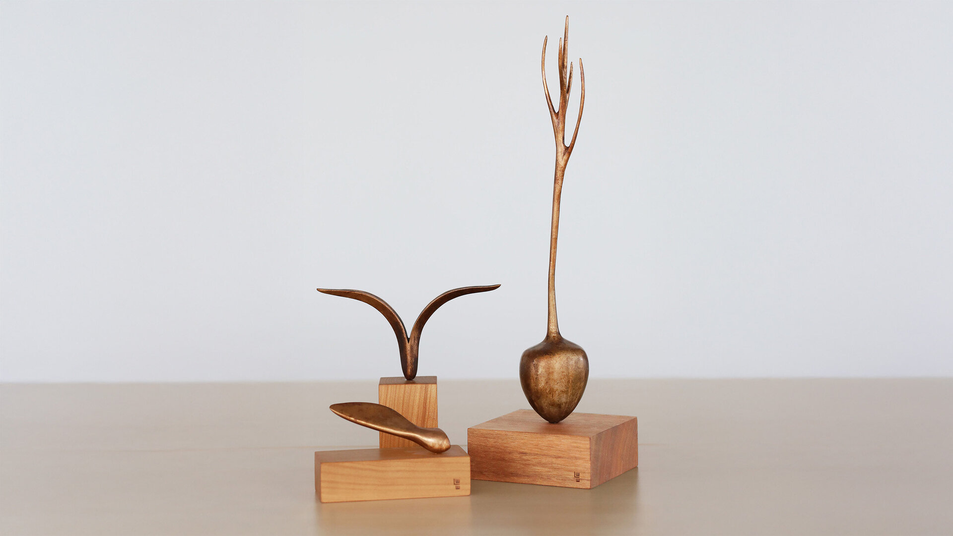 Drei filigrane Bronze-Skulpturen auf Holzsockeln vor hellem Hintergrund: eine flügelartige Form, ein tropfenförmiges Objekt und eine hohe, verzweigte Figur. Minimalistisch arrangiert, mit klarem, modernem Ausdruck.