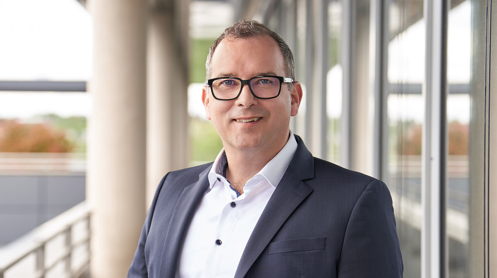 Foto von Alexander Weiss, Ressortleiter Personal und Kommunikation, stehend in Anzug und Hemd vor einem modernen Bürogebäude.