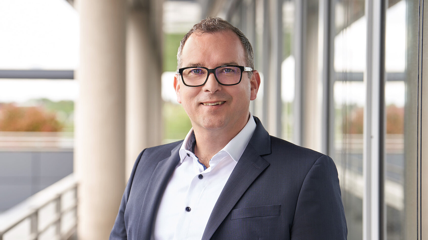 Photo of Alexander Weiss, Head of Human Resources and Communications, standing in a suit and shirt in front of a modern office building.