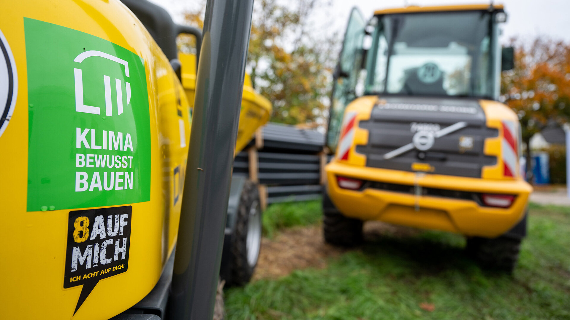 Im Vordergrund ist ein gelbes Baufahrzeug mit dem Focus auf einen Aufkleber, der auf Klimaschutz hinweist, während ein weiteres Fahrzeug im Hintergrund sichtbar ist.