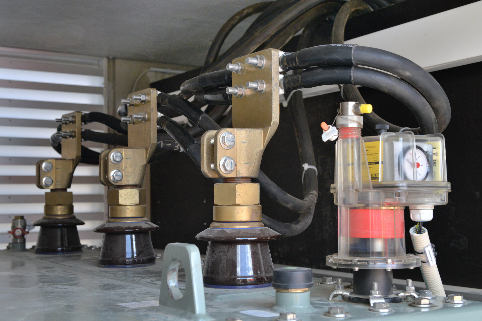 Nahaufnahme einer technischen Anlage mit mehreren dicken schwarzen Kabeln, die über große Metallklemmen an keramischen Isolatoren befestigt sind. Rechts im Bild ist ein transparenter Kunststoffbehälter mit rotem Bauteil und Manometer zu sehen. Industrielle Elektro- oder Energietechnik.