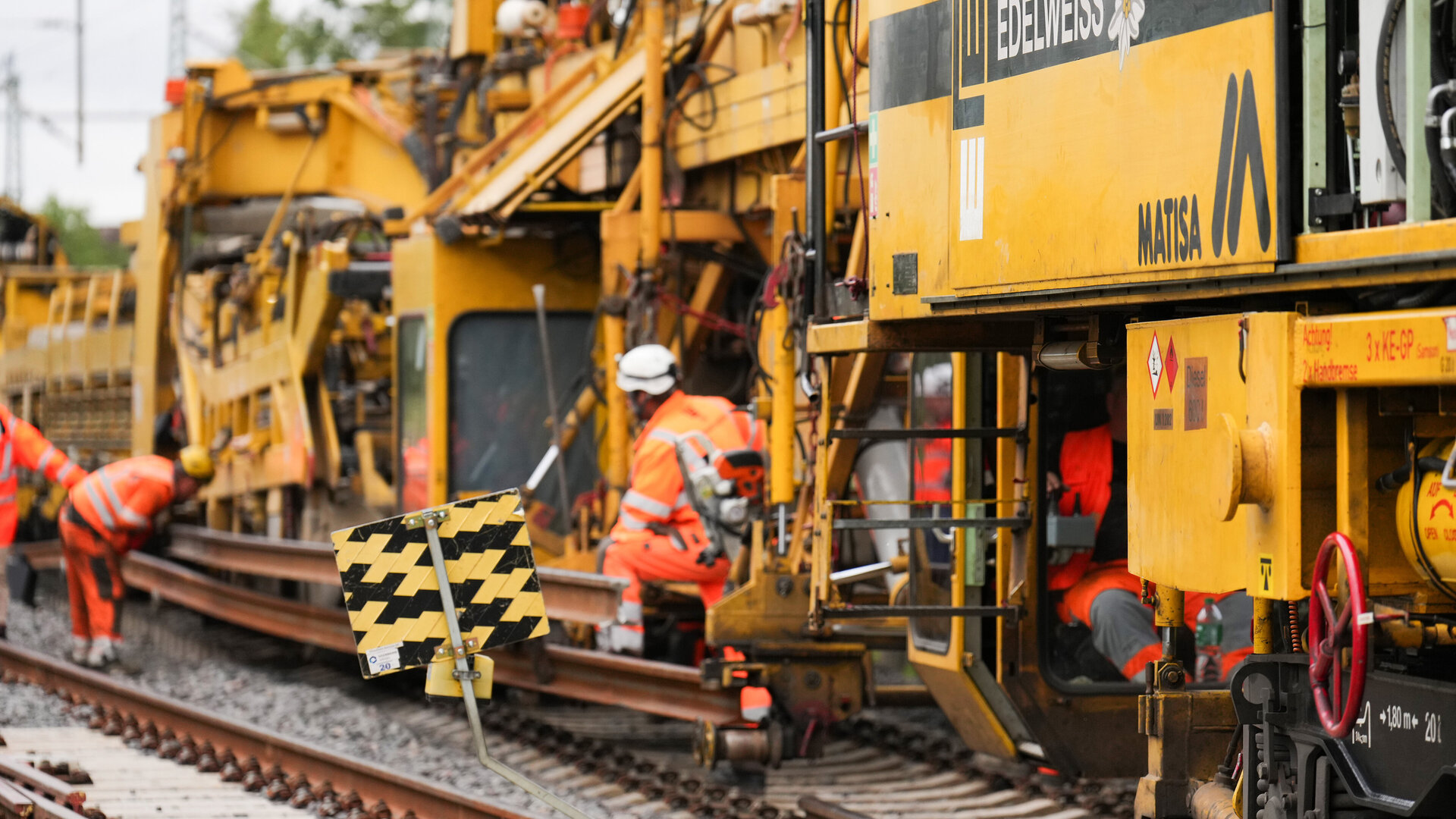 Arbeiter in leuchtend orangefarbener Kleidung führen Wartungsarbeiten an einer Bahngleisanlage mit schweren Maschinen durch.