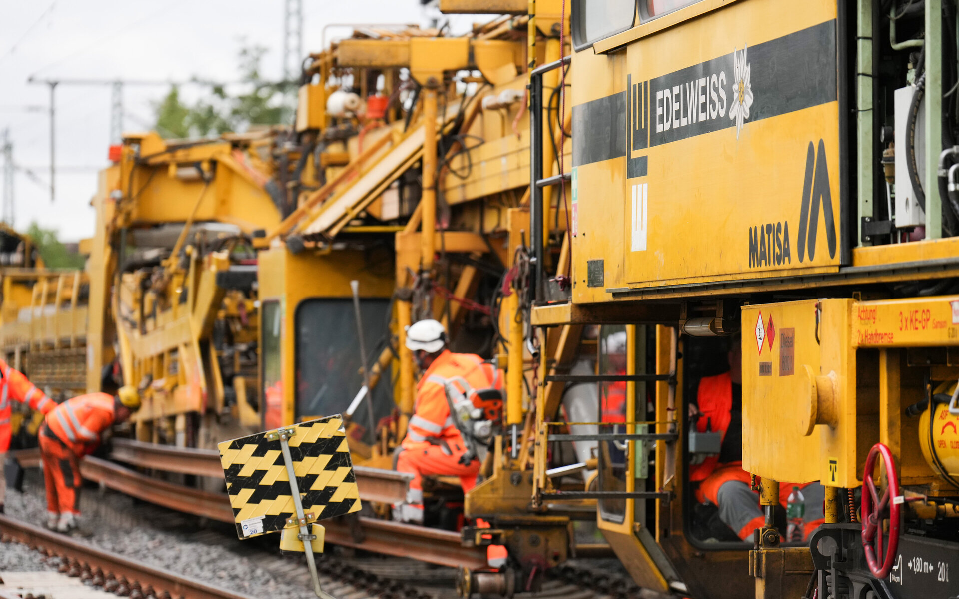 Arbeiter in leuchtend orangefarbener Kleidung führen Wartungsarbeiten an einer Bahngleisanlage mit schweren Maschinen durch.