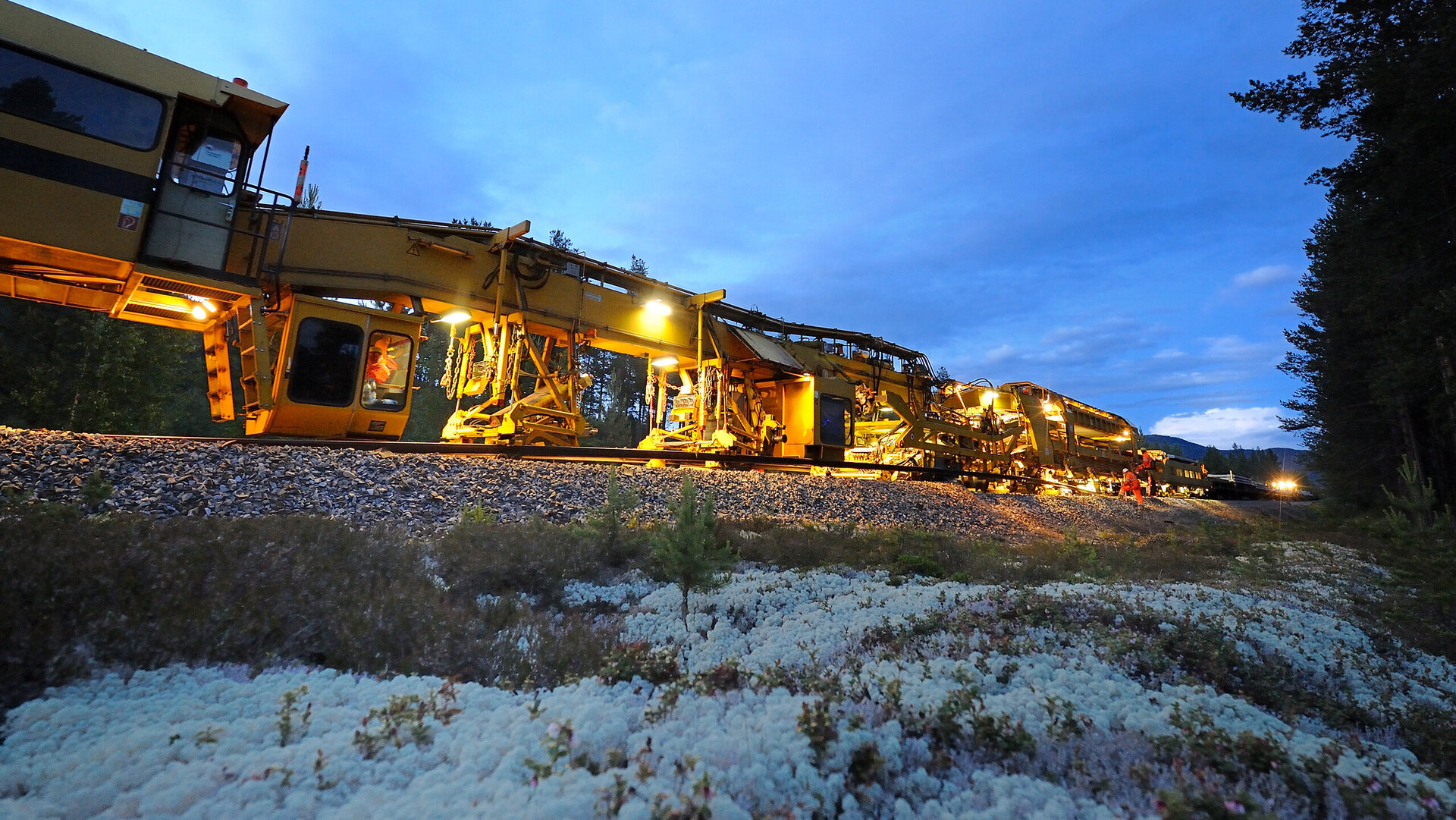 At dusk, a yellow track-laying machine can be seen working on the tracks with headlights, surrounded by nature.