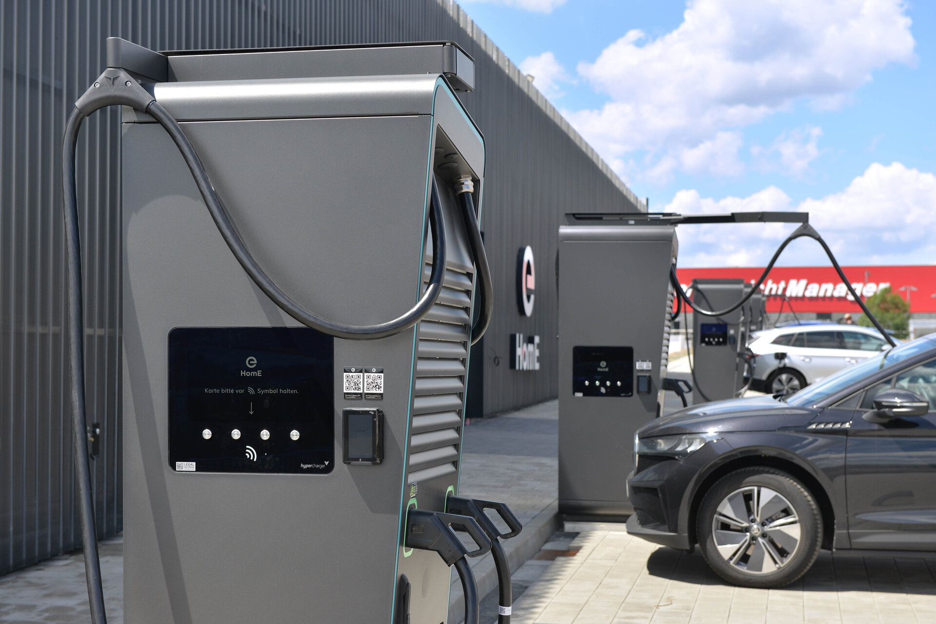 Zwei moderne Ladestationen für Elektroautos stehen auf einem Parkplatz, während ein graues Auto in der Nähe parkt.